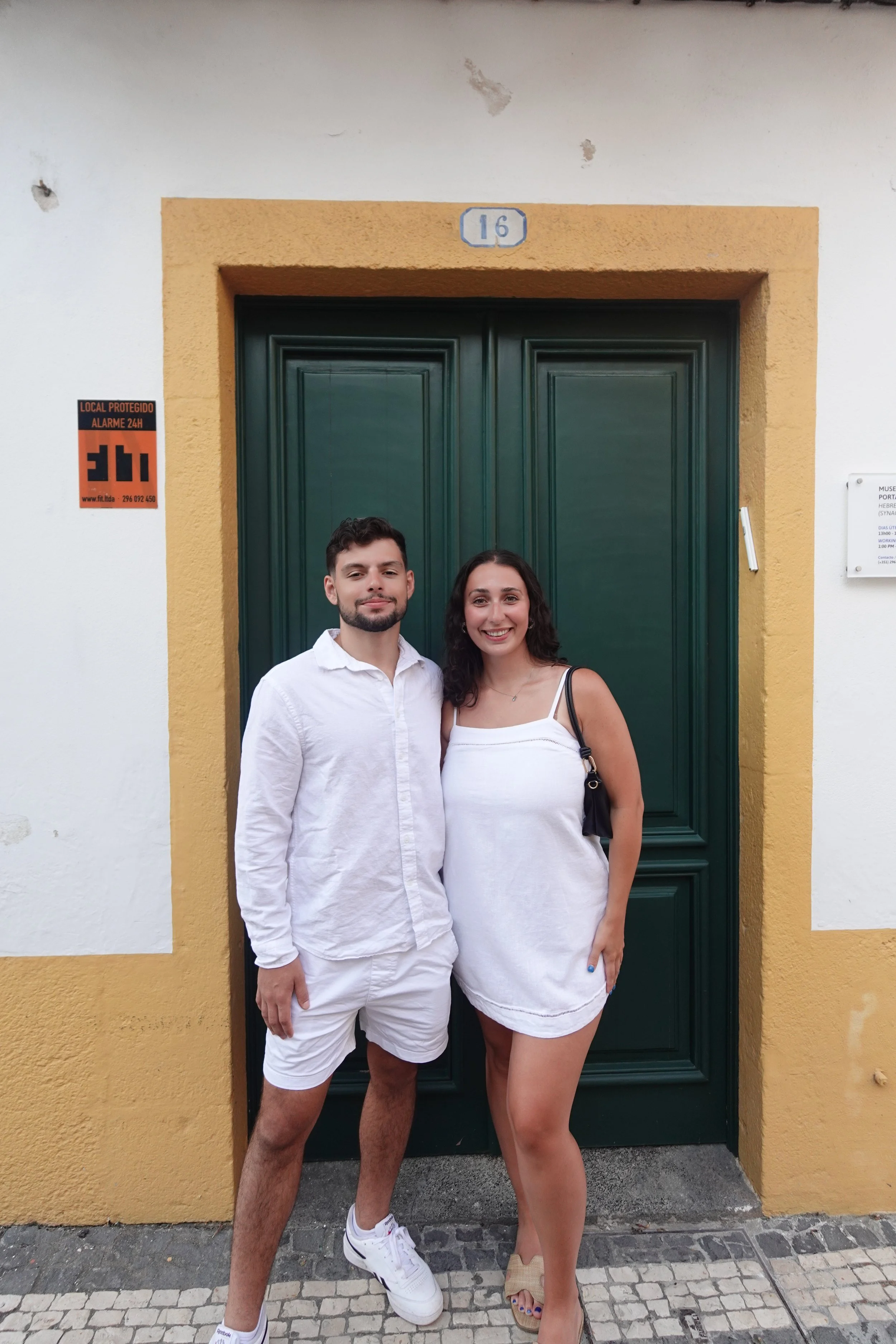 A man and woman standing close together in front of a green door with a yellow frame, smiling at the camera. The man is wearing a white shirt and white shorts, while the woman is dressed in a white sleeveless dress. They are outdoors, on a cobblestone sidewalk.