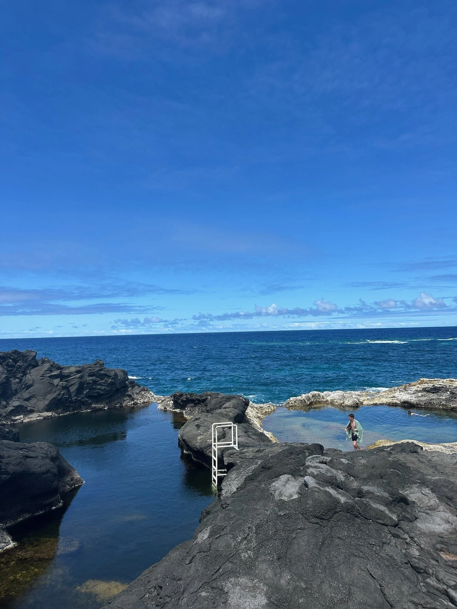A coastal scene with black volcanic rocks forming tide pools, a ladder leading into one of the pools, and a woman walking near the water. The ocean extends to the horizon under a blue sky with some clouds.