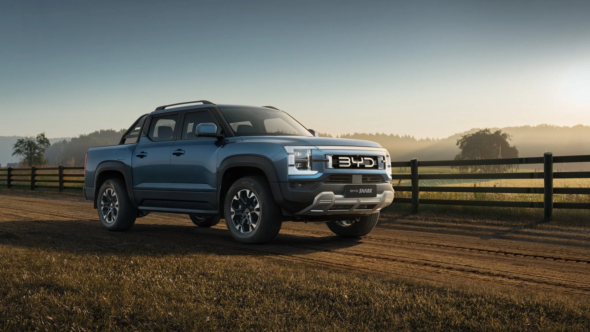 Blue BYD Shark pickup truck parked on dirt road with wooden fence and trees in the background during sunset.
