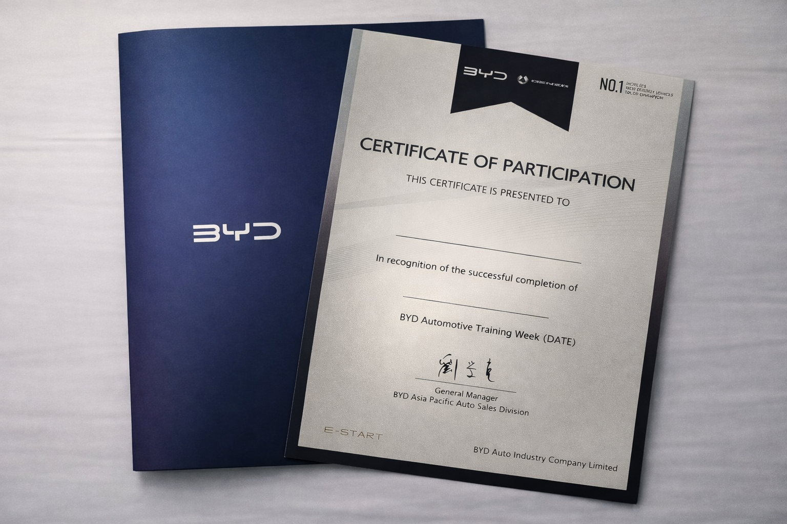 A printed certificate of participation on a silver background and a blue folder with the BYD logo, related to an automotive training week.