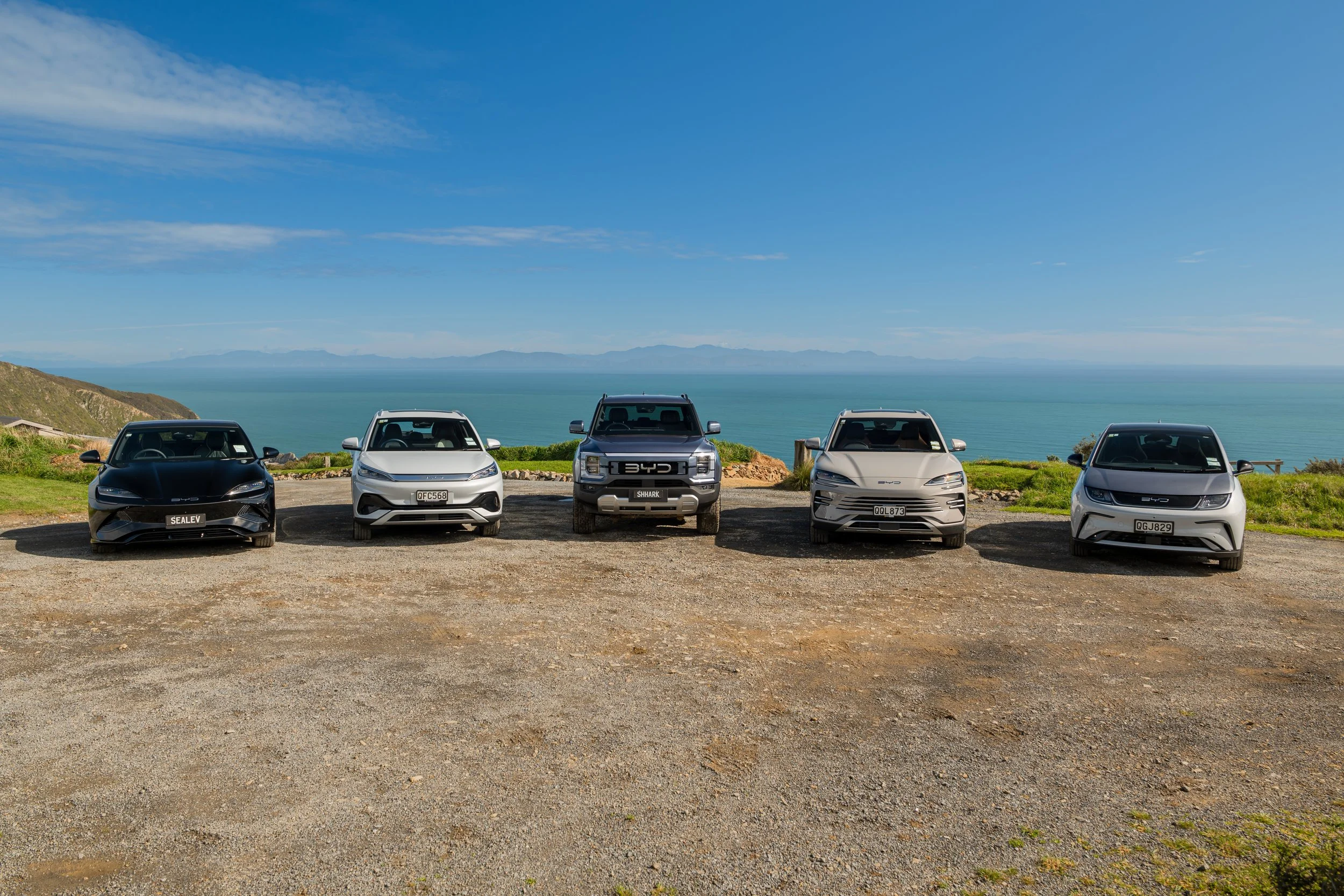 Five cars parked in a row on a dirt area with grass, overlooking the ocean and distant mountains under a clear blue sky.