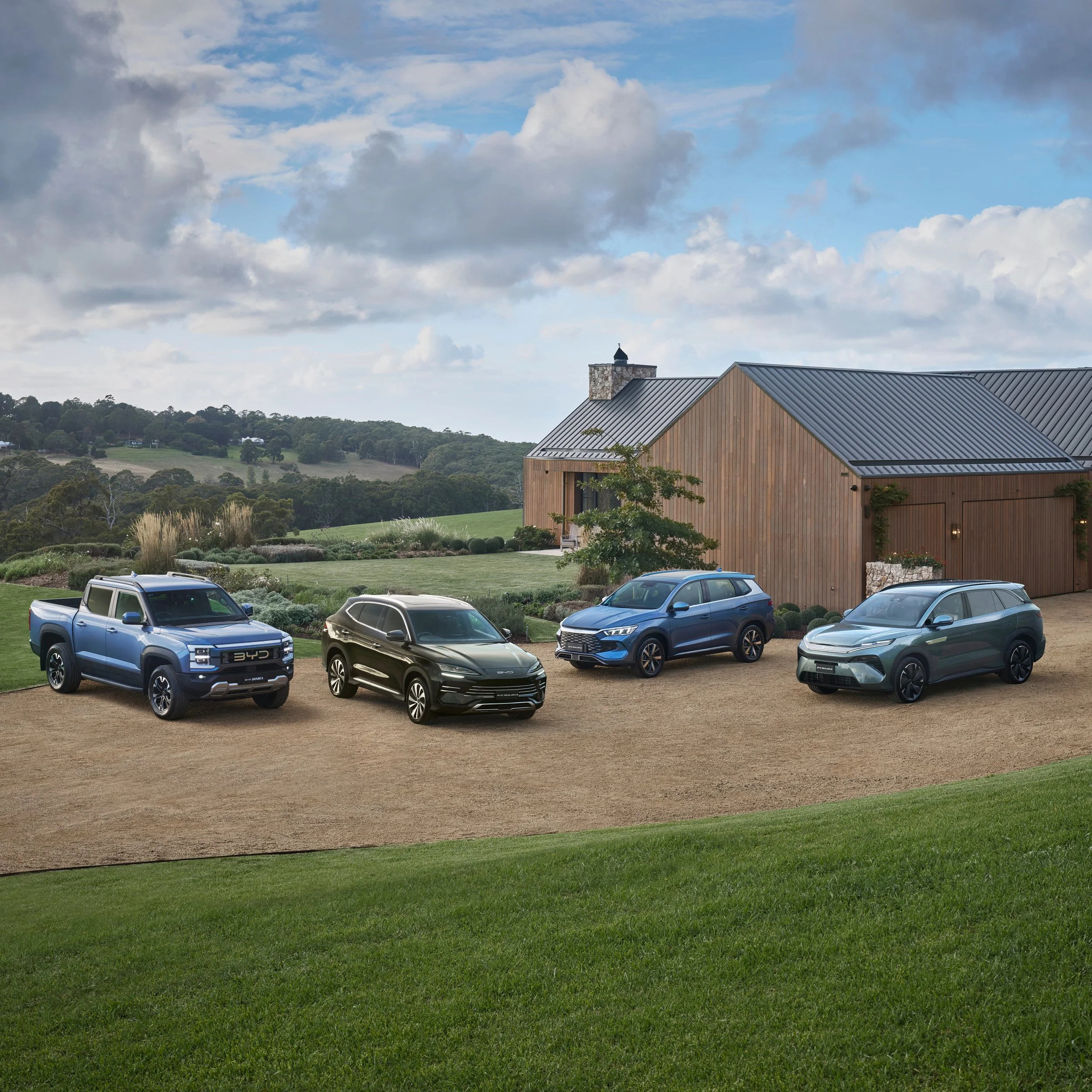 A modern house with wooden siding and metal roof, surrounded by a well-maintained lawn and parked cars, with rolling hills and trees in the background under a partly cloudy sky.