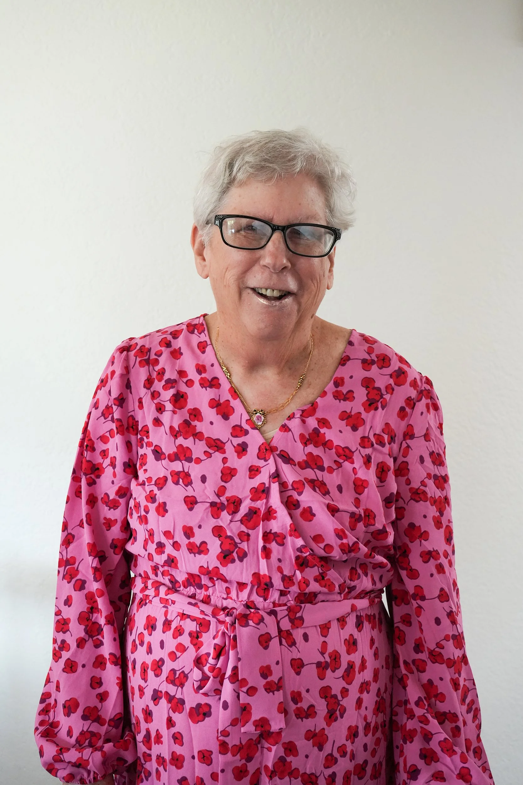 Older woman poses for photo against a blank wall in a pink dress