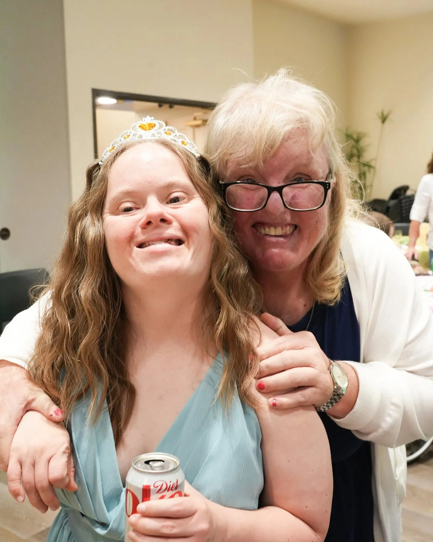 Two women posing closely in a cheerful moment, one wearing a tiara and holding a can of Diet Coke, both smiling widely.
