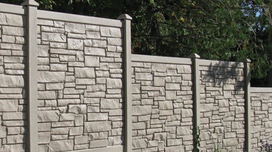 A tall, decorative concrete fence designed to look like stacked stone, with pillars dividing each section. Trees are visible behind it.