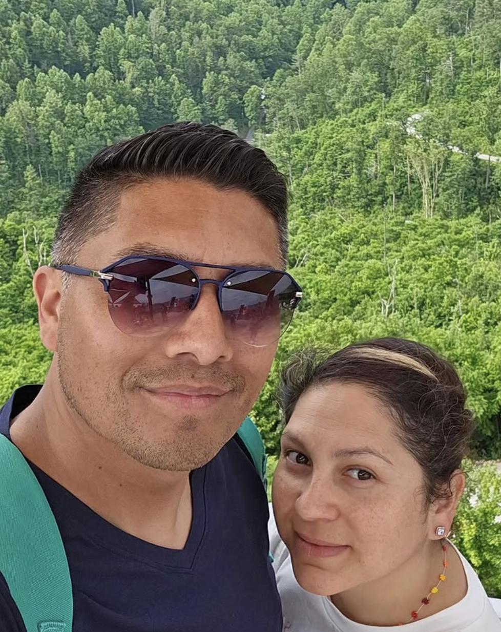 A man and woman taking a selfie outdoors with a lush green forest in the background.