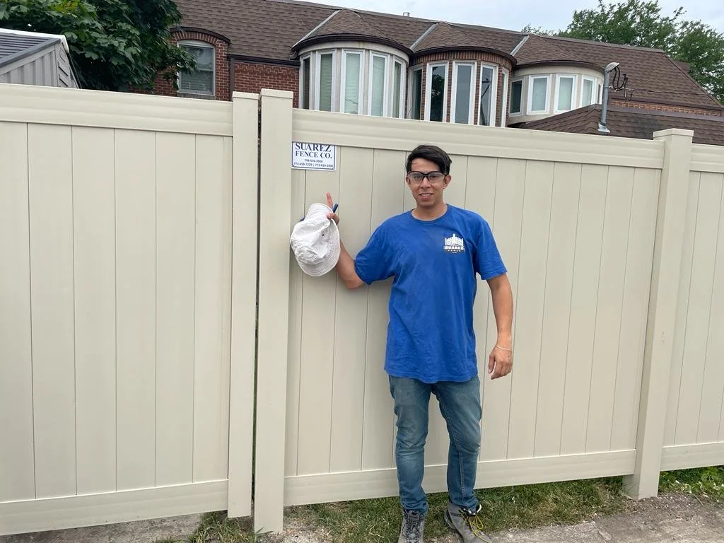 Man standing next to a beige vinyl privacy fence, giving a thumbs-up, holding a white cap, with a sign that reads 'Suarez Fence Co.' in a residential neighborhood.