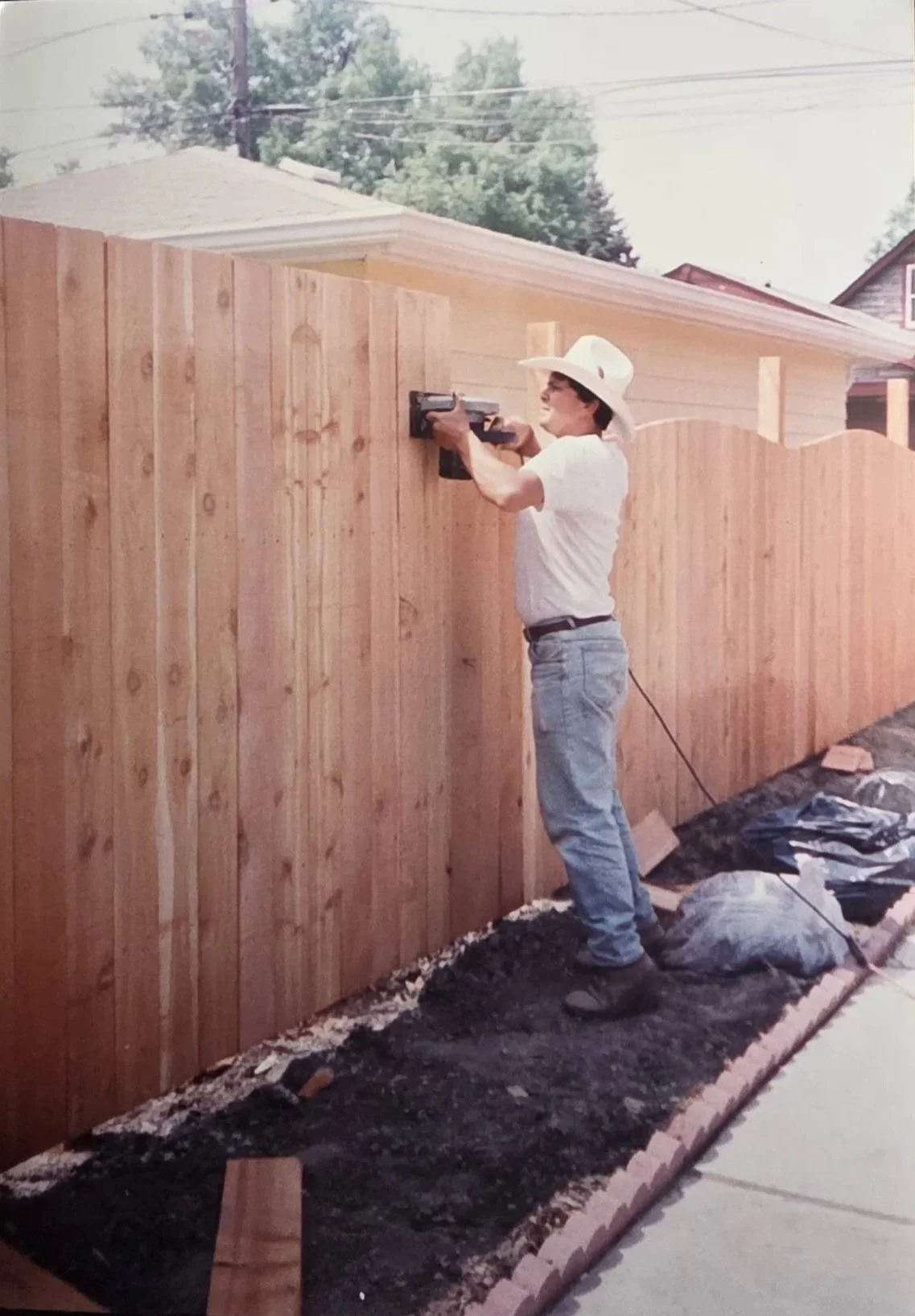 A person in jeans and a cowboy hat uses a power tool to build a wooden fence in a yard on a sunny day.