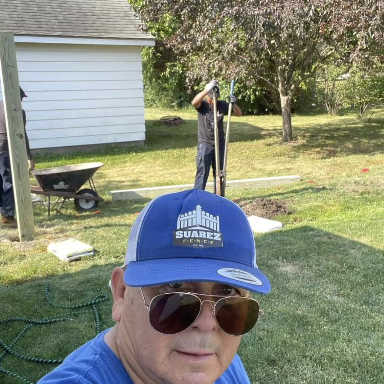 A man wearing a blue cap with 'Suarez Fence' logo, sunglasses, and a blue shirt taking a selfie in a backyard where a person is working on installing a fence, with a white shed, green grass, and trees in the background.