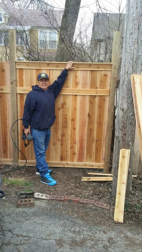 Man in a navy hoodie and jeans, wearing a cap, standing in front of a wooden fence under construction, holding a power drill in his right hand, outdoors with residential houses and trees in the background.