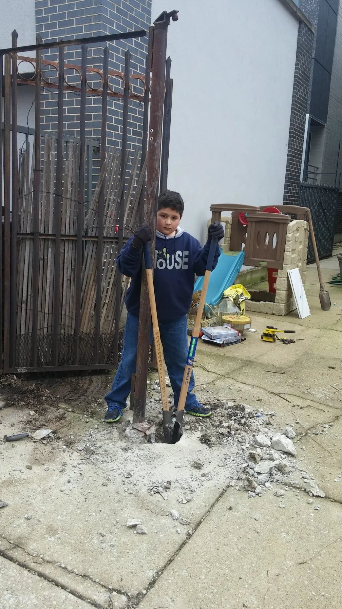 A young boy standing outside on a concrete surface, holding a small shovel and a pickaxe, with some construction debris around a hole in the ground.