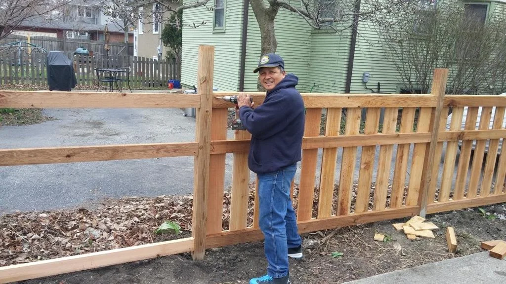 A man in a blue hoodie and jeans installing a wooden fence with a power drill in a backyard.