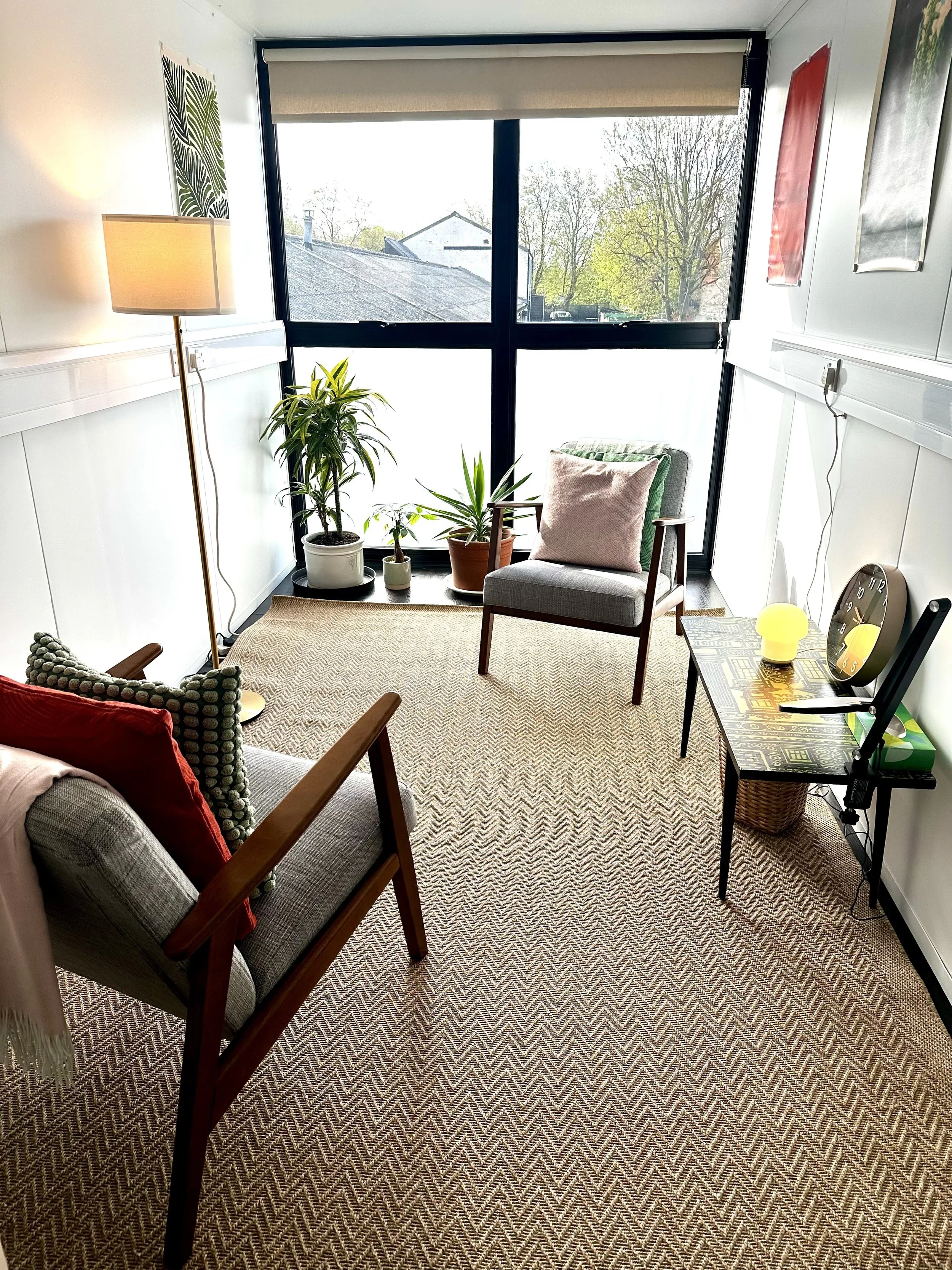 Cozy sunlit sitting room with a large window, two armchairs with pillows, indoor plants near the window, a side table with a clock and a lamp, a floor lamp, and wall art.