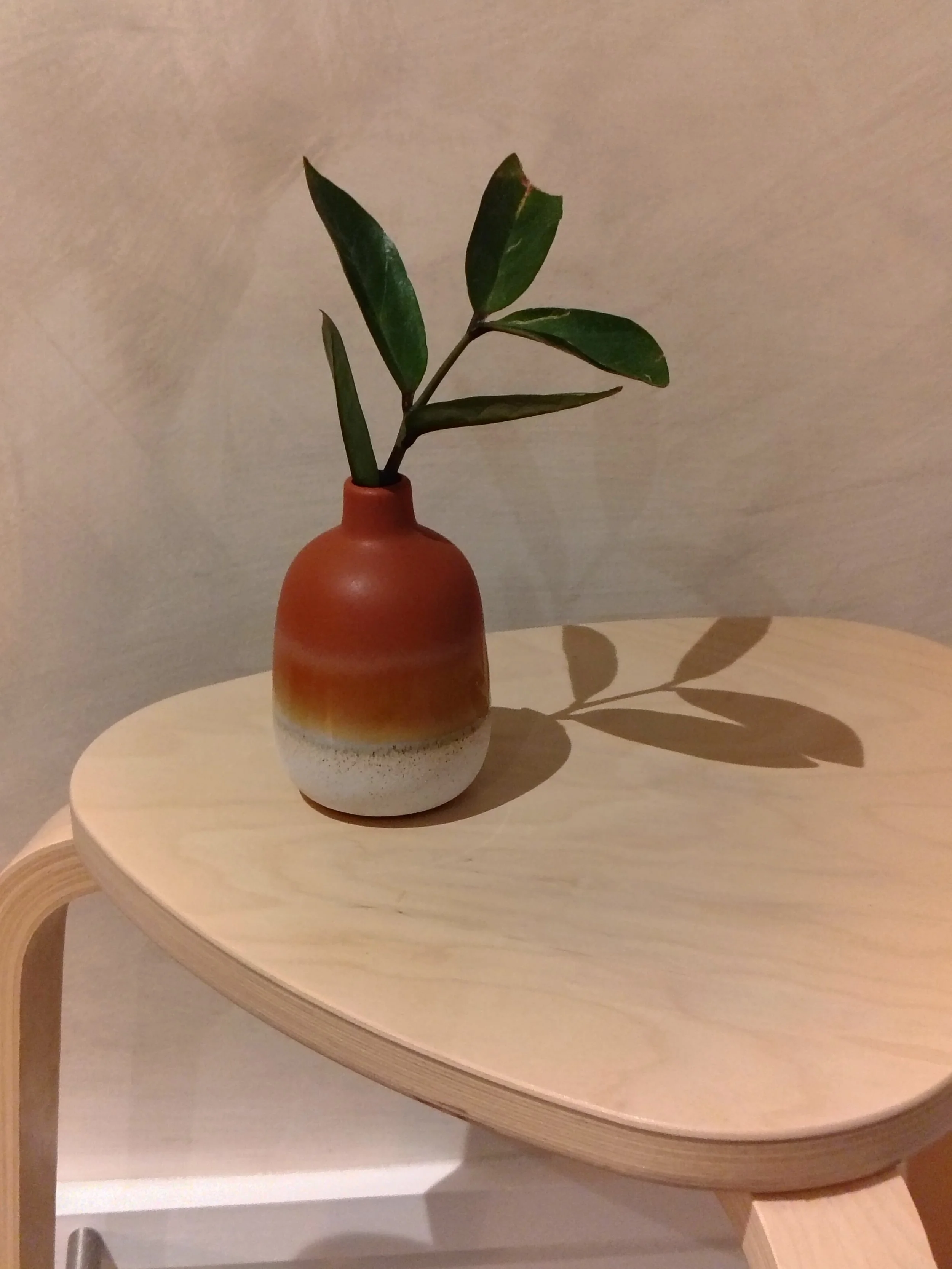 A small ceramic vase with a gradient color from brown to white, holding a green leafy branch, placed on a light-colored wooden table with rounded edges.