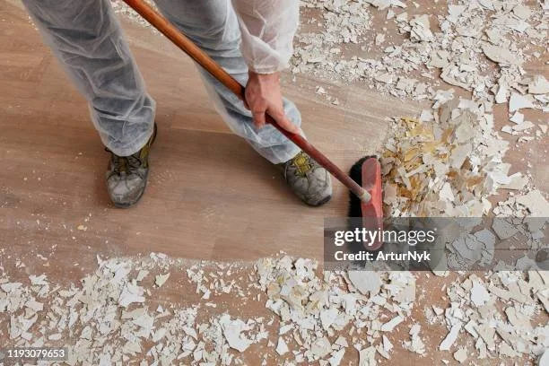 Une personne en gilet de protection qui enlève la vieille peinture d'un plancher à l'aide d'une raclette.
