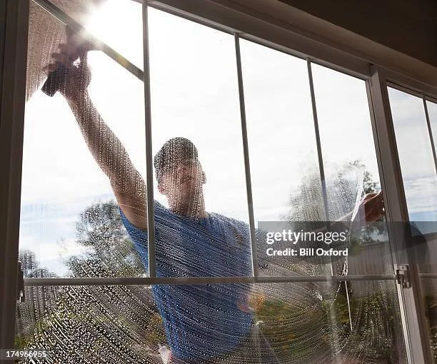 Une personne nettoie une grande vitre du dehors avec un chiffon, vue de l'intérieur d'une pièce.