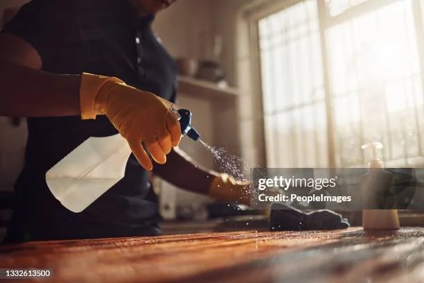 Une personne portant des gants en caoutchouc utilise un spray de nettoyage sur une surface en bois dans une cuisine.