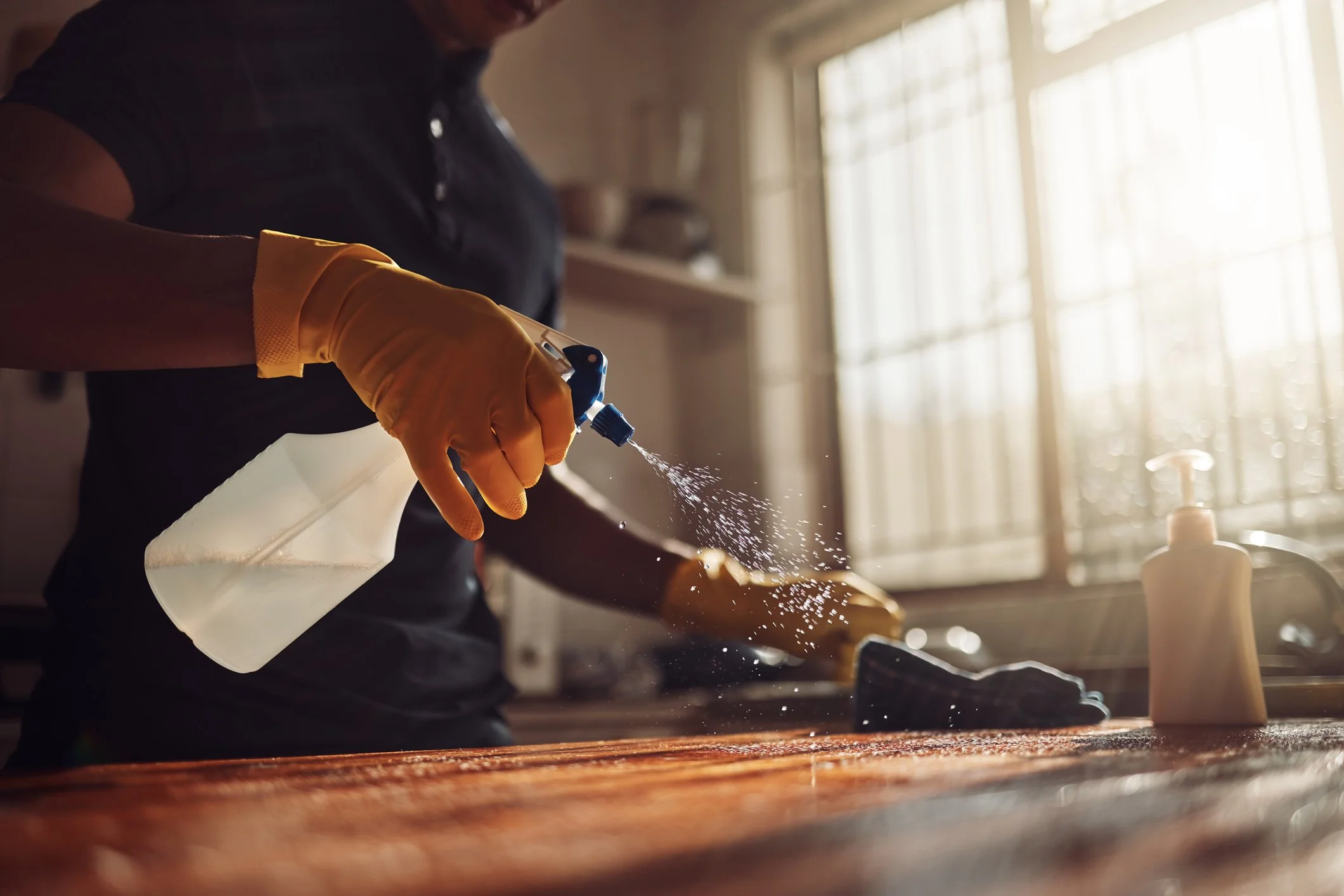 Une personne portant des gants en caoutchouc utilise un spray de nettoyage sur une surface en bois dans une cuisine.