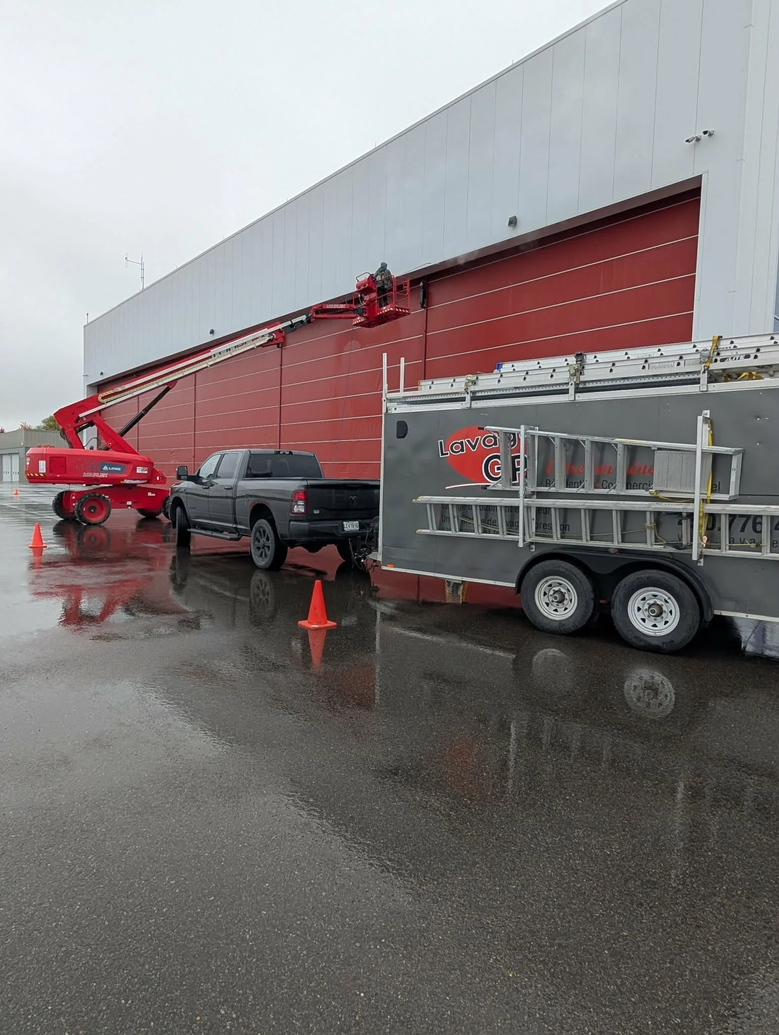 Transporteur utilisant une nacelle élévatrice pour effectuer des travaux sur une grande porte de hangar rouge, avec un véhicule noir et une remorque à côté, sous un ciel nuageux par temps de pluie.