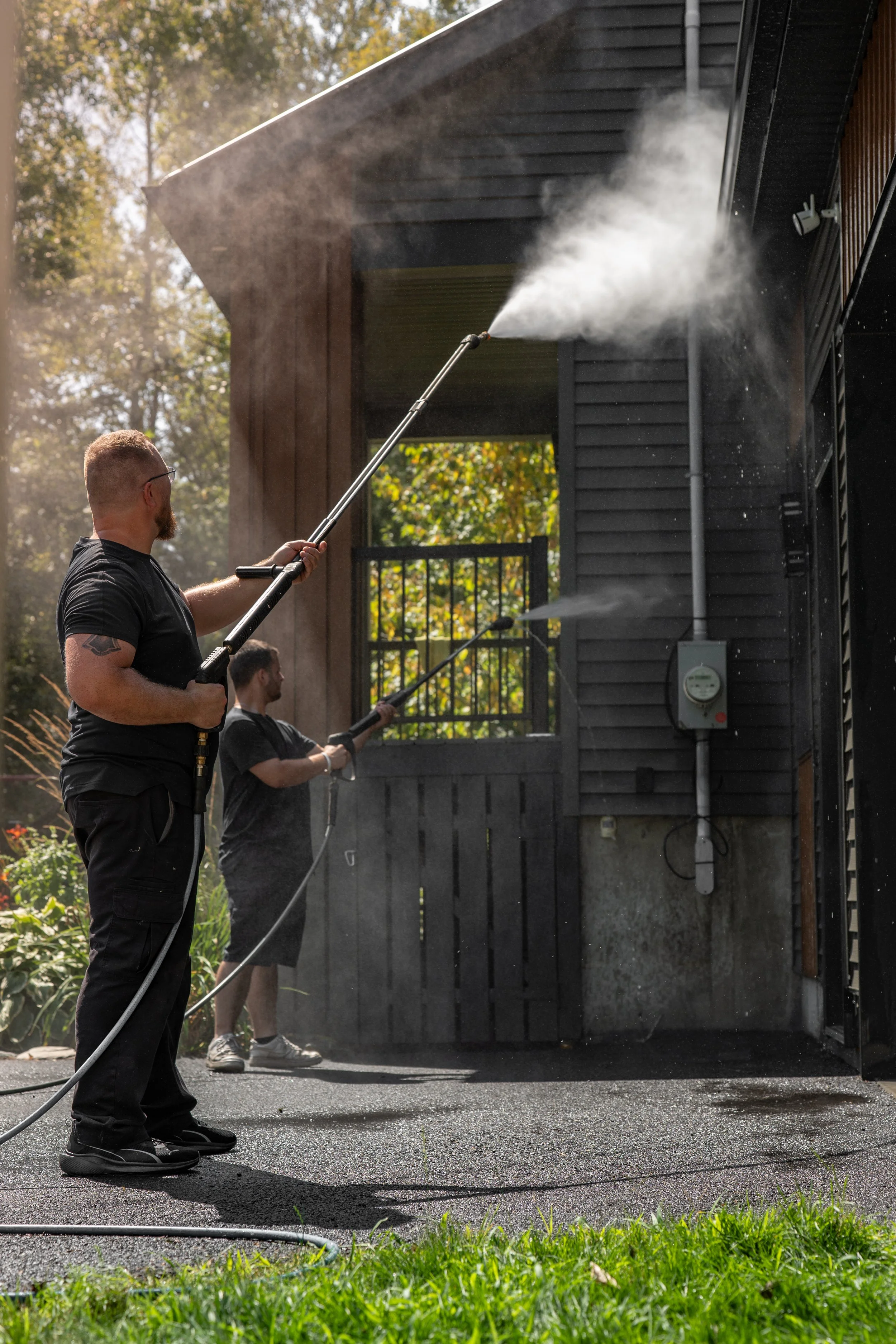 Deux hommes utilisent des tuyaux haute pression pour nettoyer la façade d'une maison en bois noir, avec un jardin en arrière-plan.