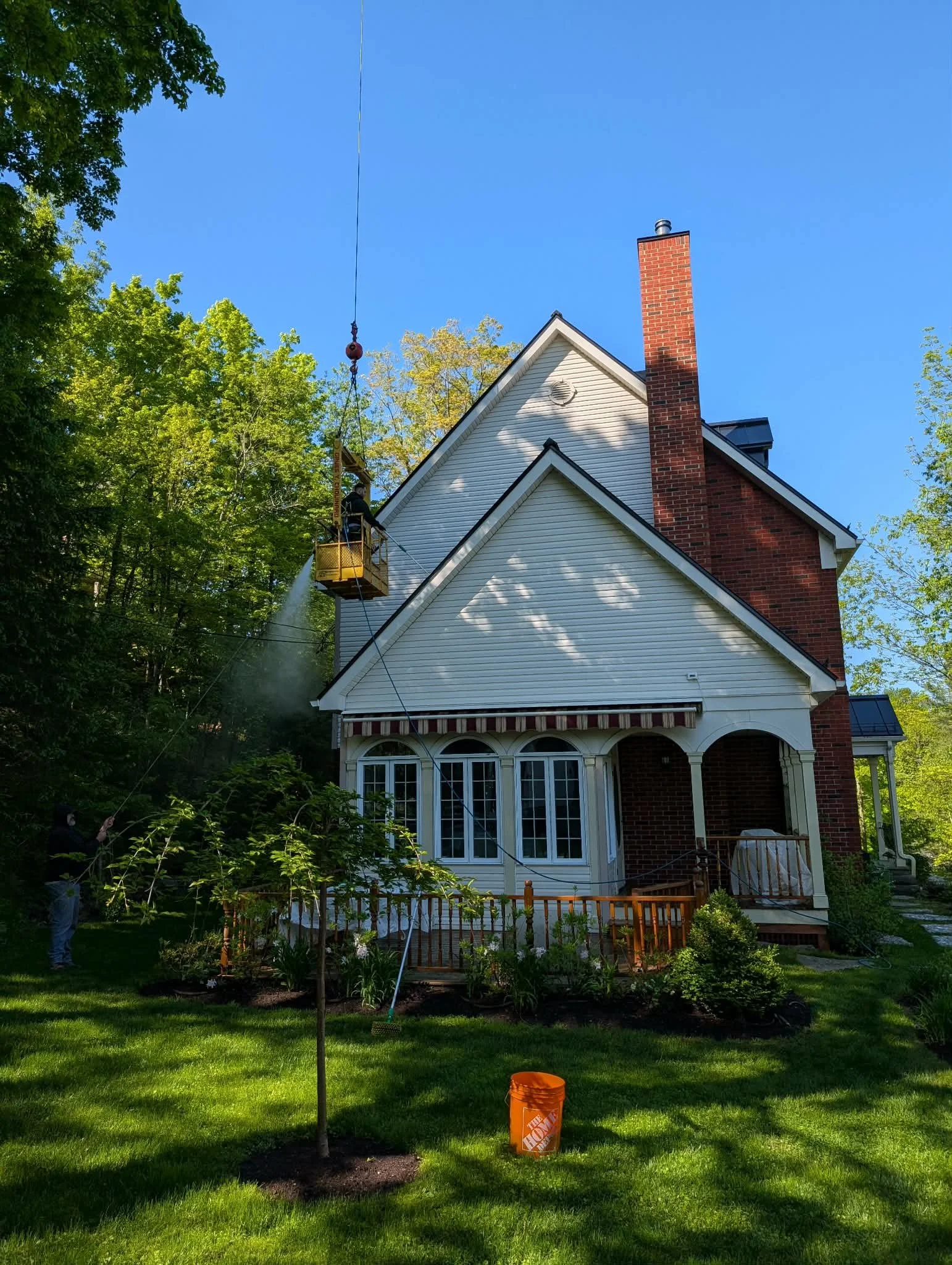 Une maison avec un jardin où deux personnes nettoient la toiture à l'aide d'une nacelle suspendue par une grue.