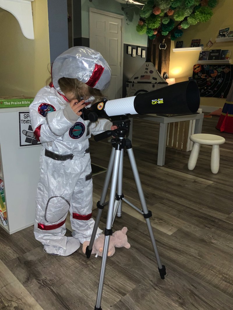 Child dressed in a white astronaut costume looking through a telescope in a room with toys and decorations.
