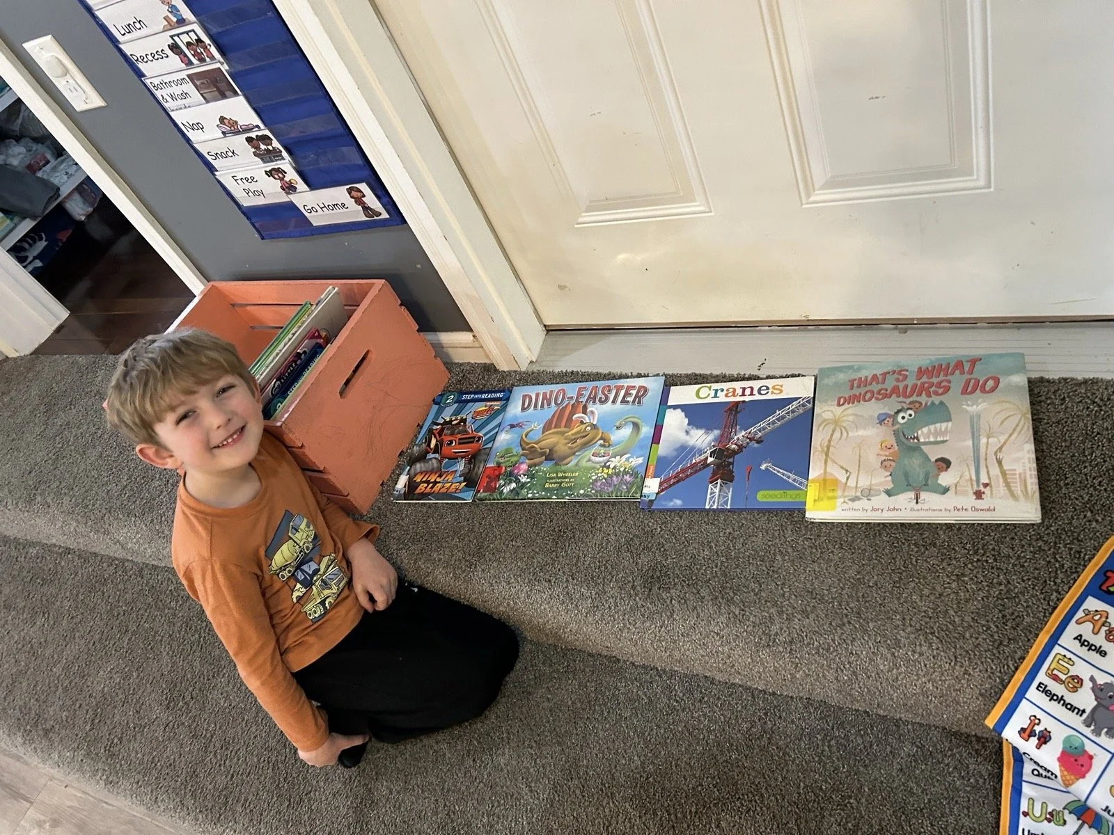 A young boy with a big smile sitting on a gray carpeted bench next to a row of children's books. The books include titles about dinosaurs, construction, and animals. There is a small orange storage box with more books nearby, and a blue and white educational poster on the wall with labels for different parts of the school day.