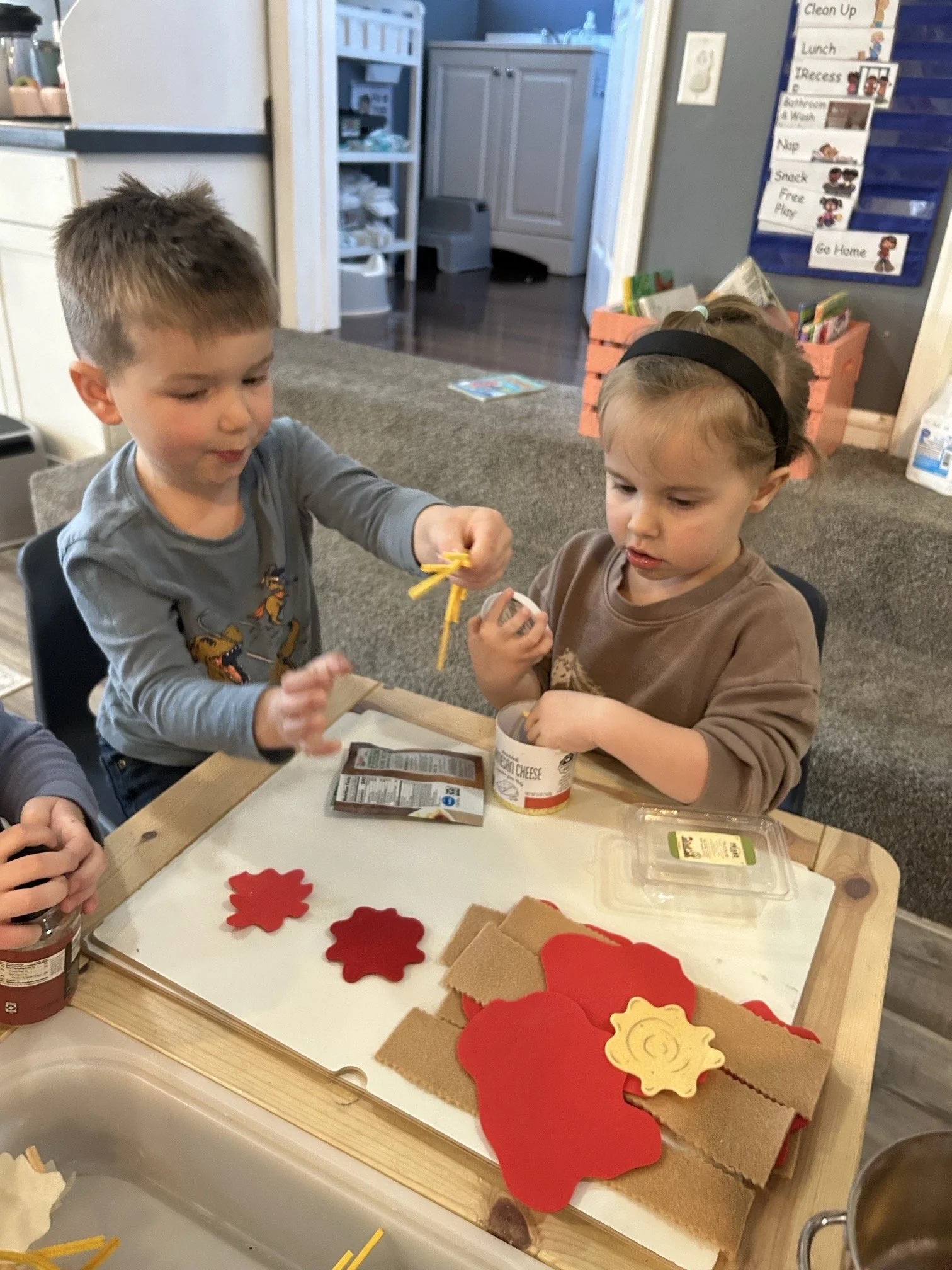 Two young children, a boy and a girl, sit at a small table in a classroom or daycare, playing with snack items and foam craft supplies, including foam shapes and string, with a background of classroom decor and storage.