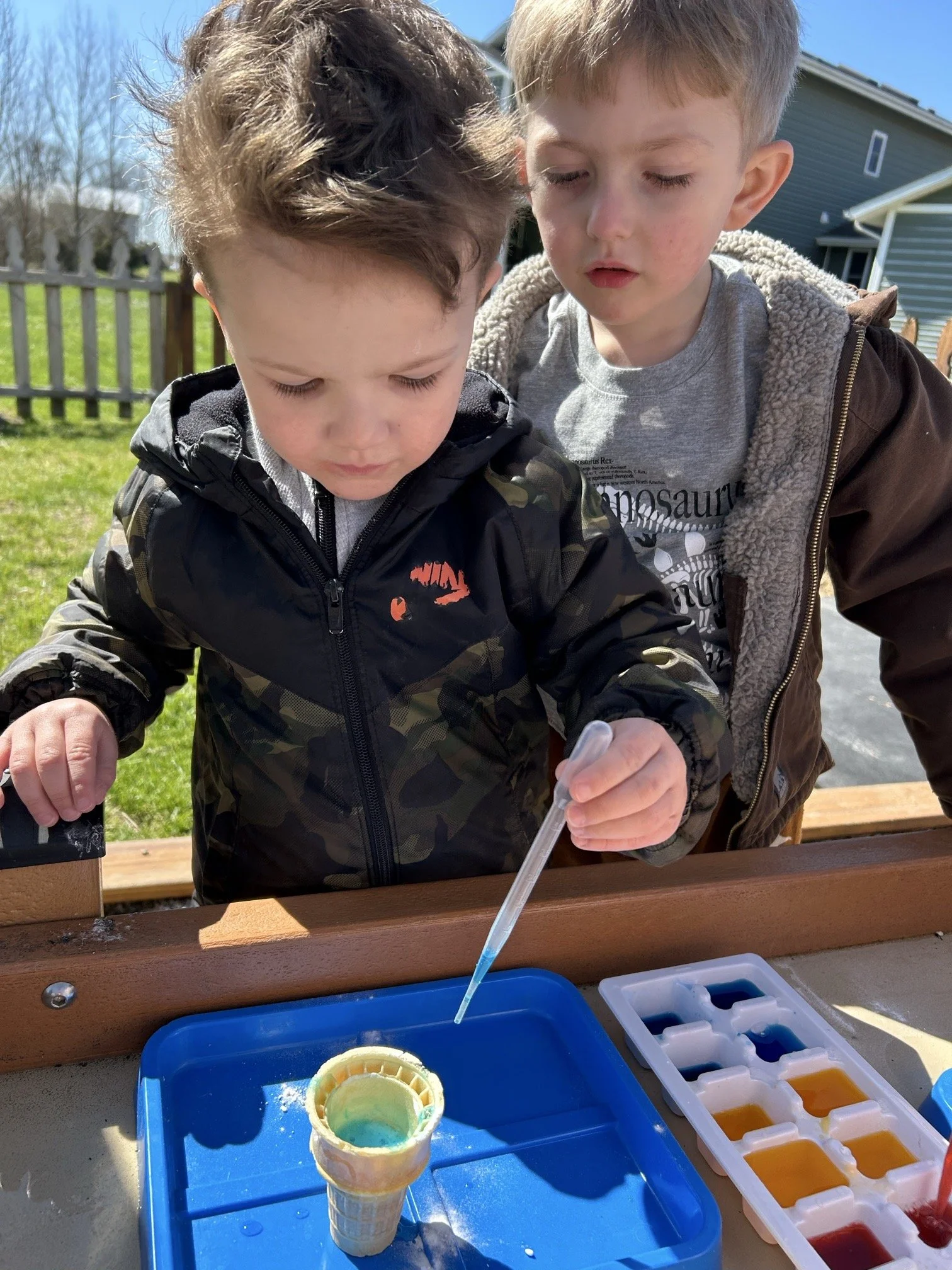 Two young boys outdoors on a sunny day, one boy with brown hair in a camouflage jacket and another with blond hair in a brown jacket, using a dropper to extract colored liquid from a container into an ice cream cone. There is a blue tray and an ice cube tray with colored liquids on a wooden surface, with a house and a fence in the background.