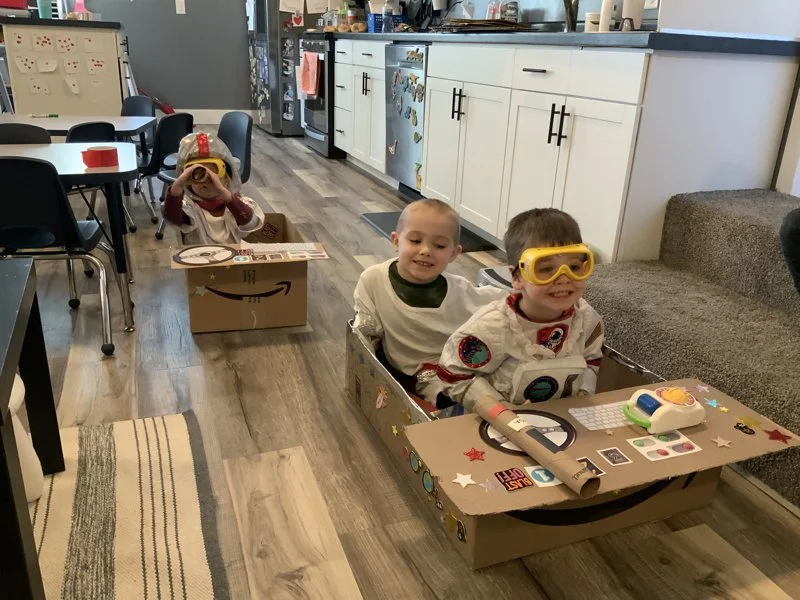 Three children in cardboard box spaceships with toy control panels, wearing astronaut costumes and goggles inside a home kitchen and living room.
