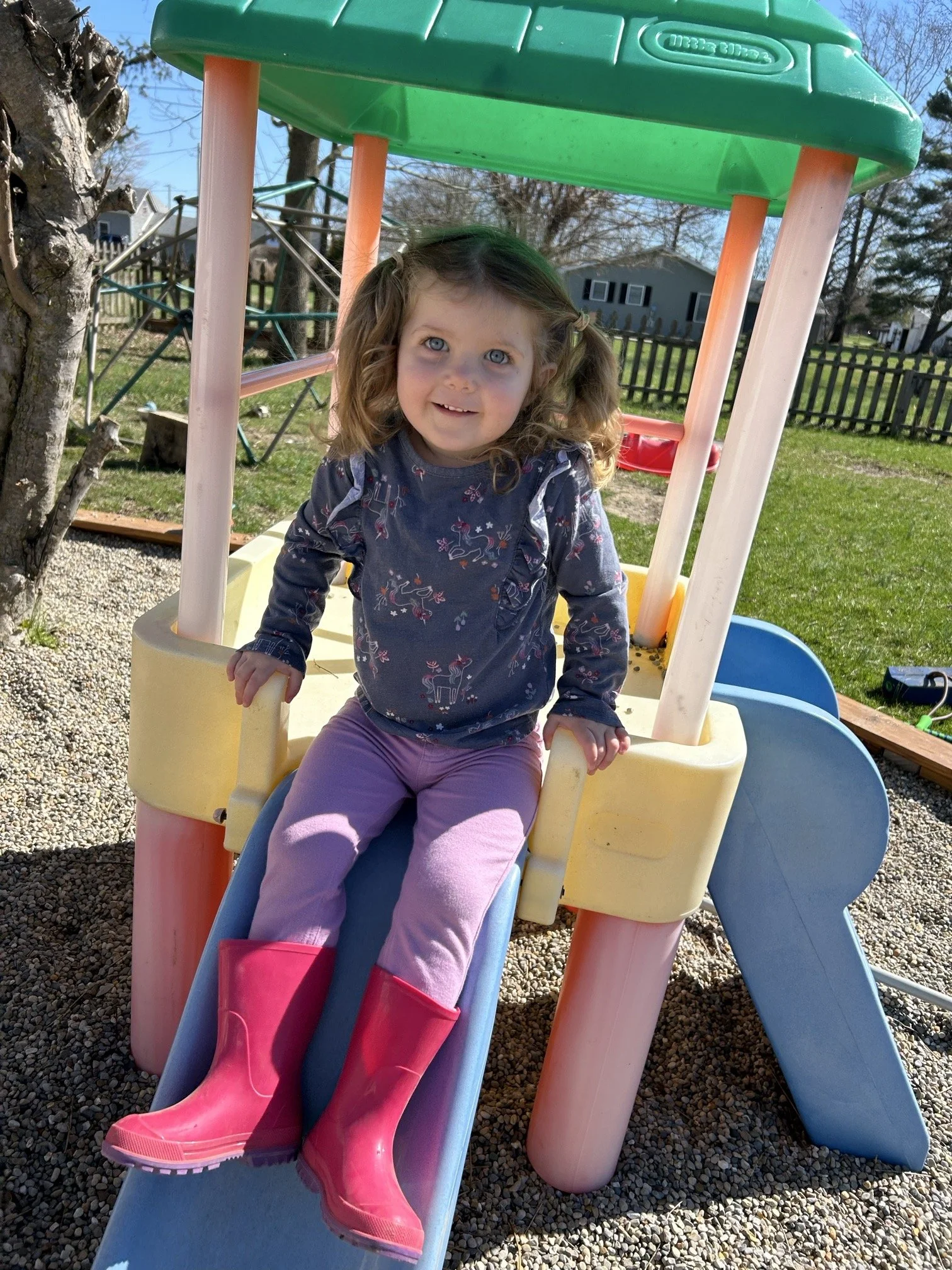 A young girl with curly blonde hair and blue eyes sitting at the top of a small plastic slide in a backyard play area, wearing a dark floral long-sleeve shirt, pink pants, and bright pink rain boots.