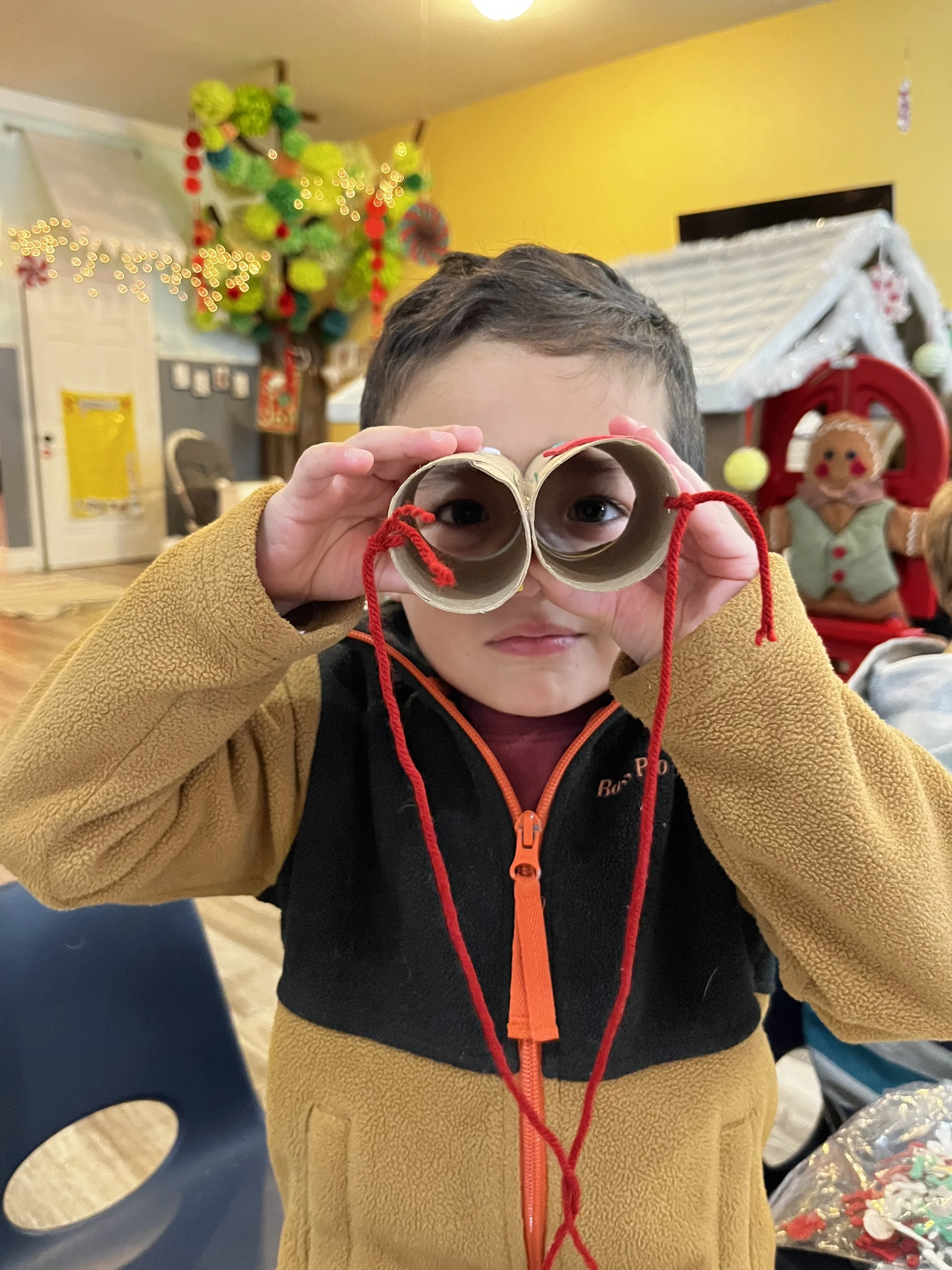 Young boy holding two rolled-up pieces of paper with red string, pretending to look through binoculars, in a festively decorated room with Christmas and holiday decorations.