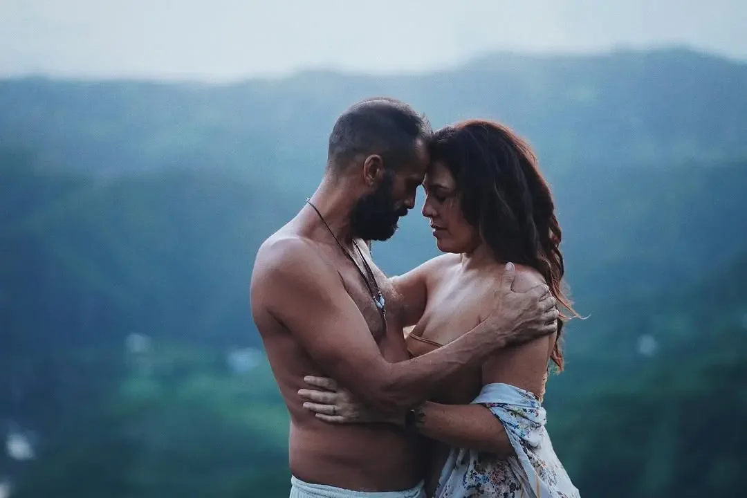 Una pareja desnuda abrazándose en un paisaje natural con colinas y cielo en el fondo.