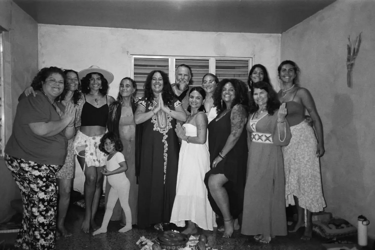 Grupo de personas posando juntas en una reunión familiar o celebración en una habitación con paredes lisas y ventanas con persianas. La escena incluye adultos y dos niñas pequeñas, algunos con vestimenta elegante y otros con ropa casual, mostrando alegría y amistad.