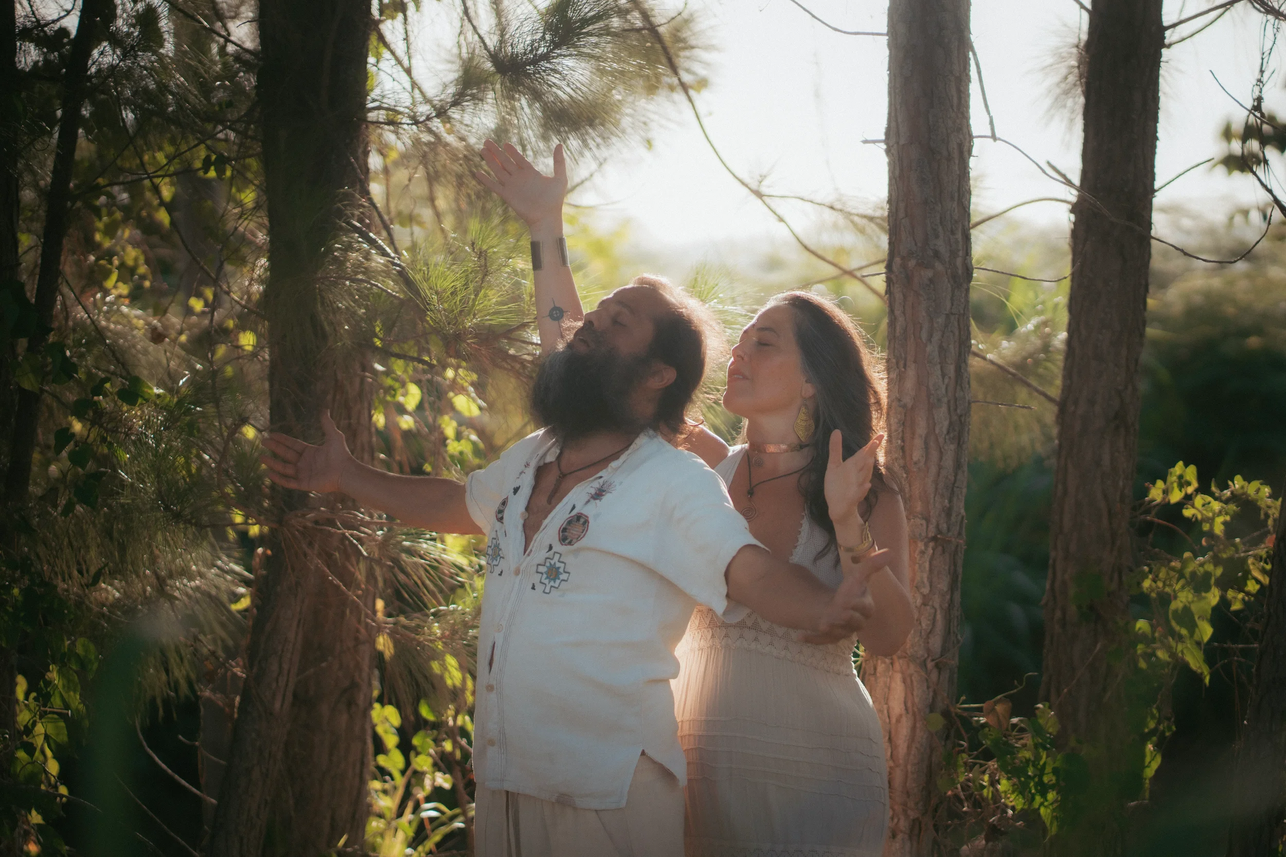 Dos personas, un hombre con barba y mujer, en un bosque, con árboles y luz solar filtrándose, en actitud de meditación o oración con los ojos cerrados.