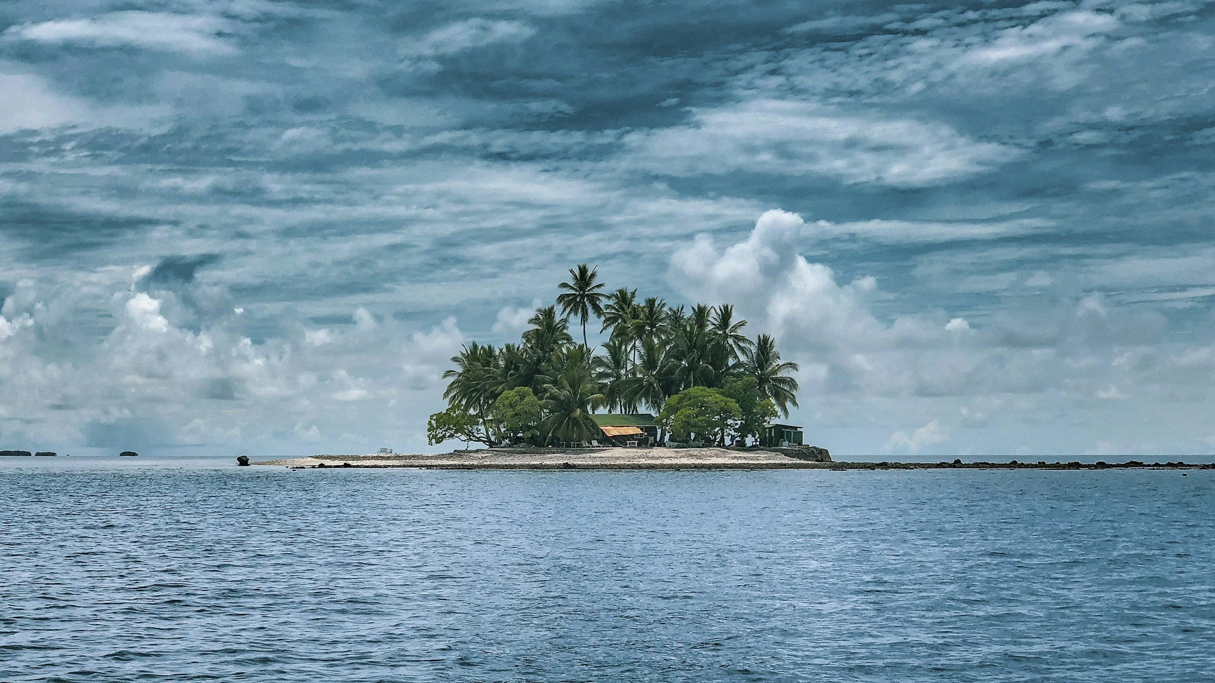 Small island with tall palm trees and a few small structures, surrounded by calm blue ocean under a partly cloudy sky.
