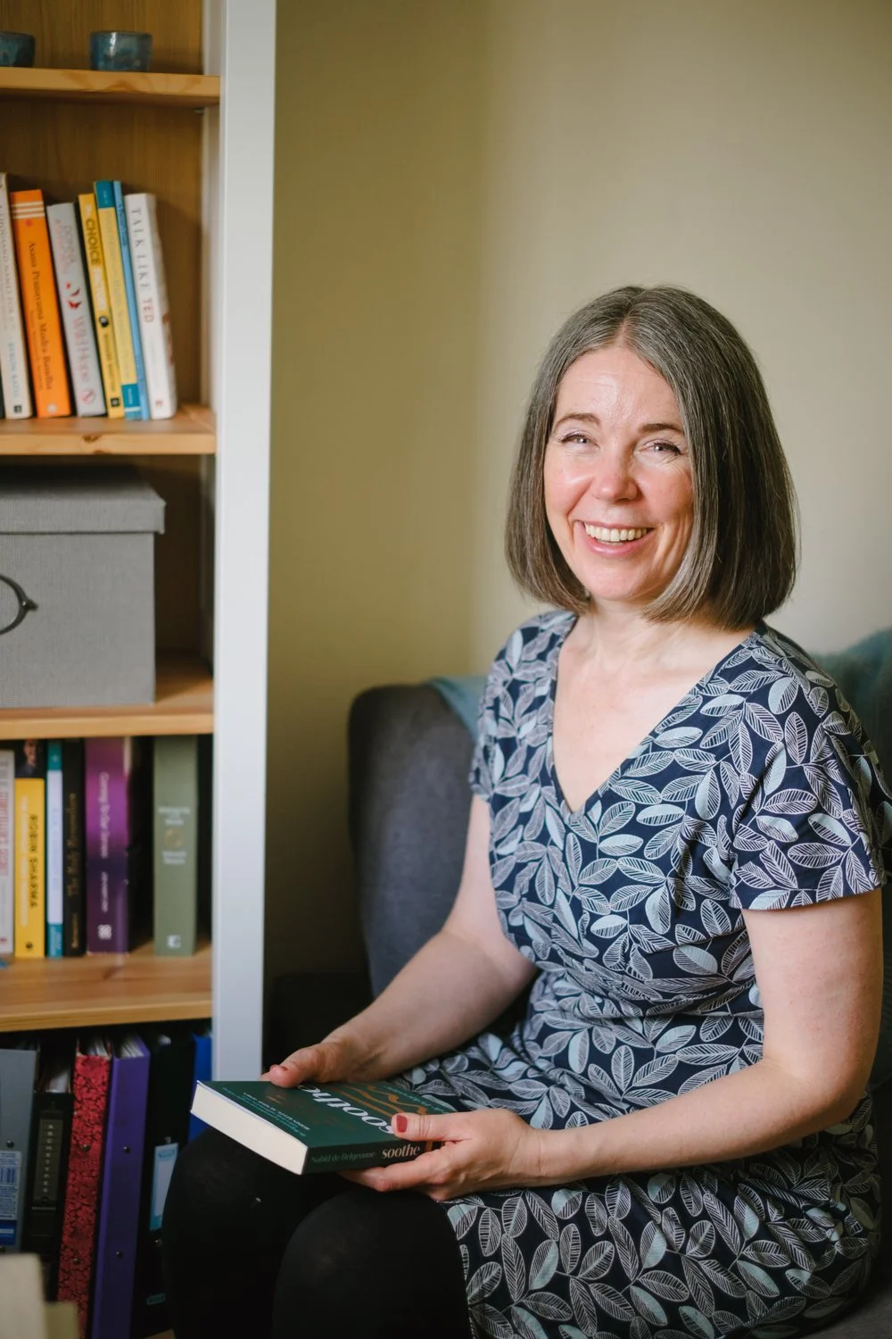 Nicola Pott, therapist, seated beside a bookshelf and holding a book, smiling warmly