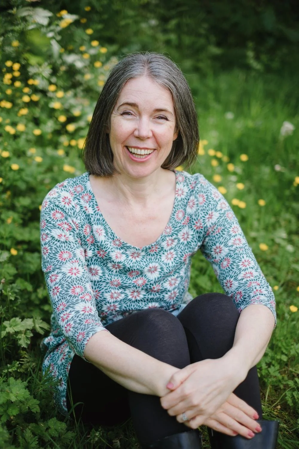 Nicola Pott, therapist, smiling while seated in a sunlit meadow of yellow wildflowers