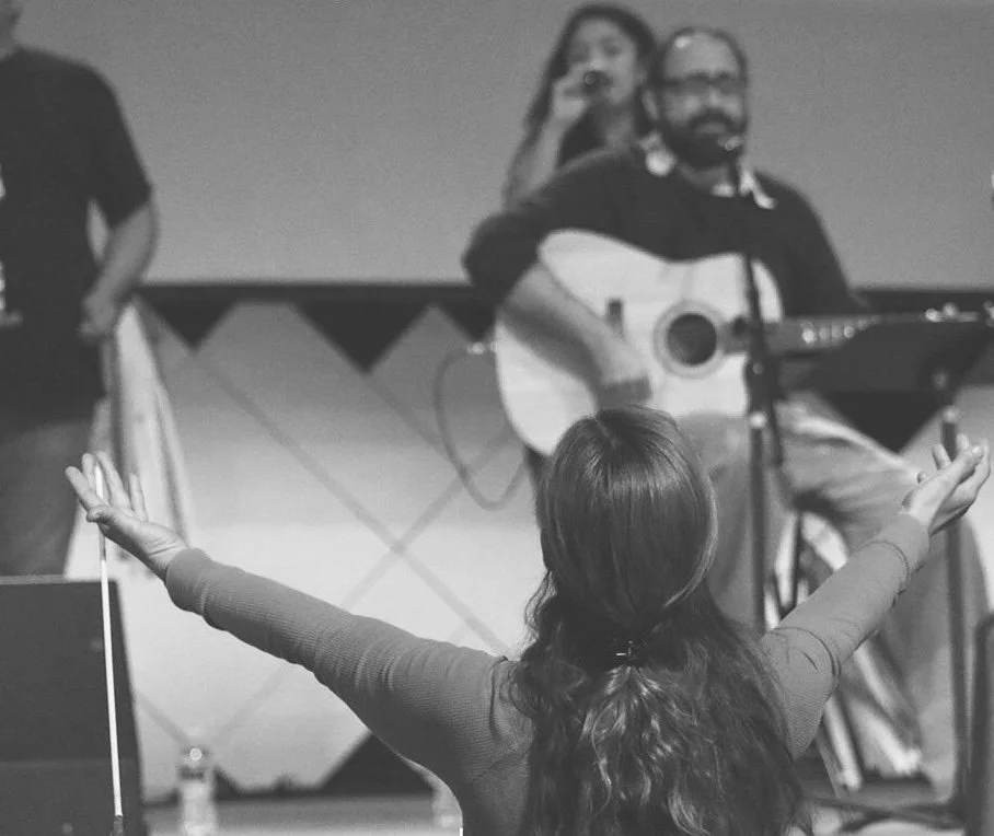 A woman with long hair and outstretched arms facing a man with glasses playing an acoustic guitar and singing, in a room with a stage and a woman in the background with a microphone.