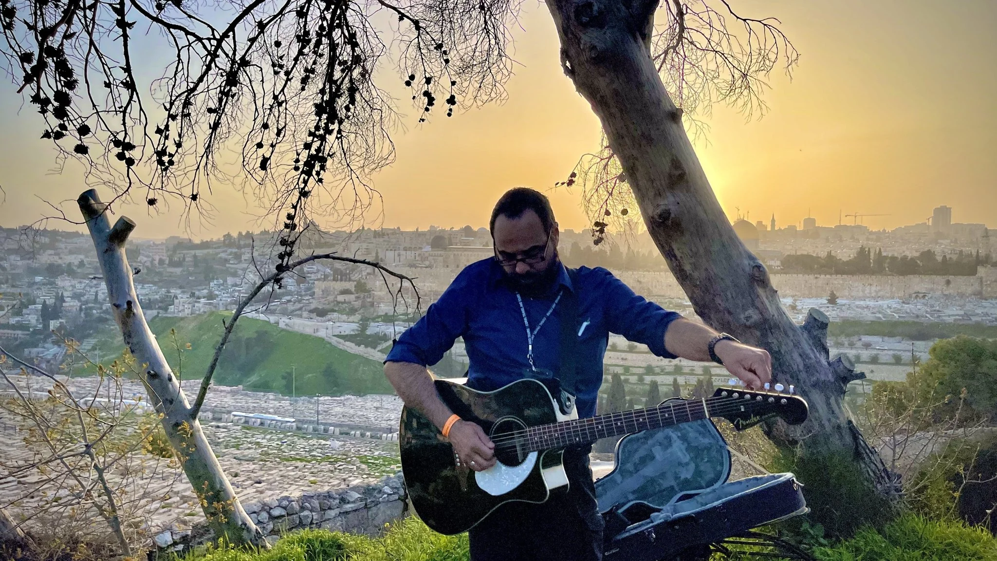 A man playing an acoustic guitar outdoors at sunset, standing near trees with a cityscape in the background.
