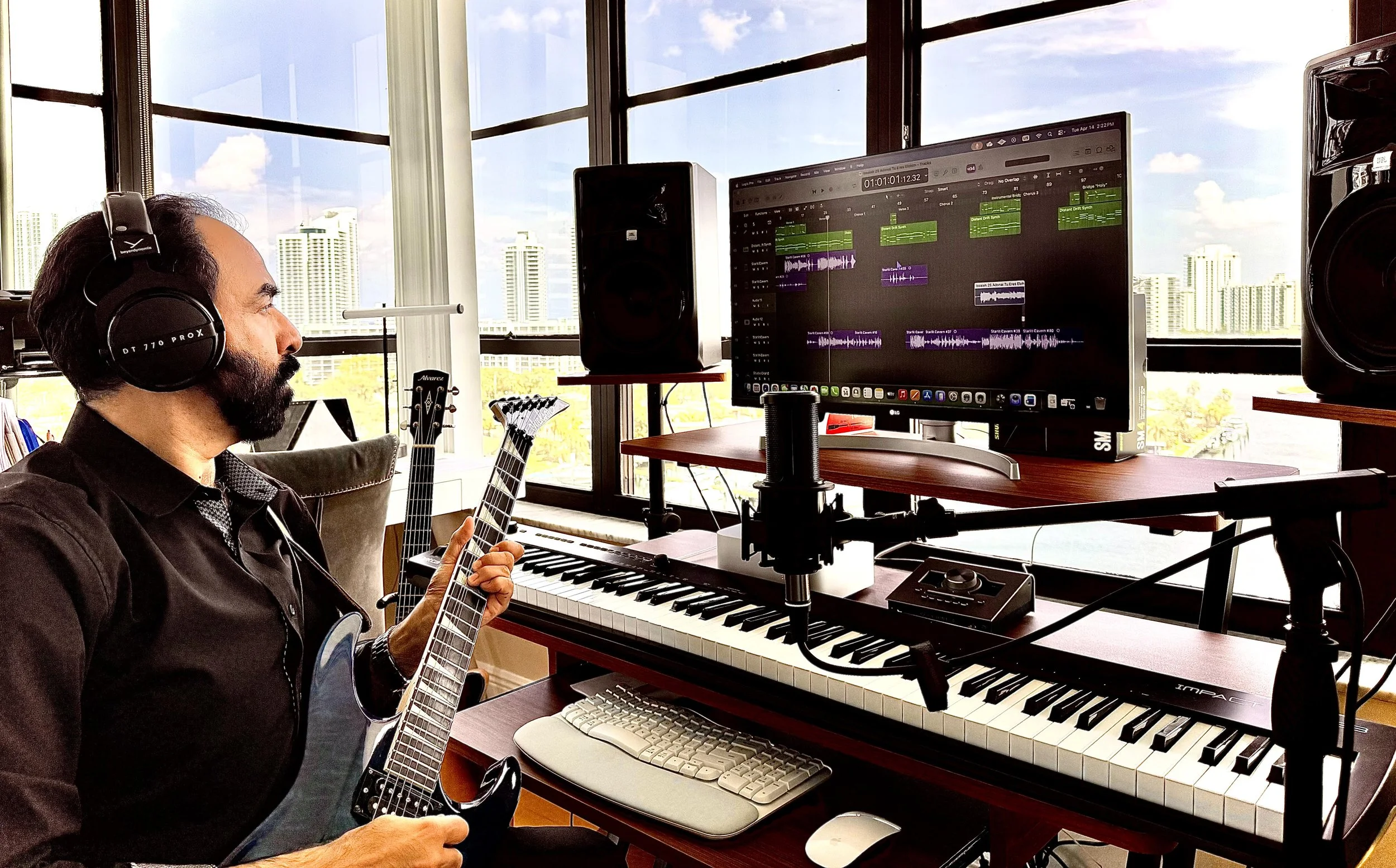 Person wearing headphones playing an electric guitar in a music studio with a large monitor, keyboard, microphones, and speakers, near windows showing a city skyline.