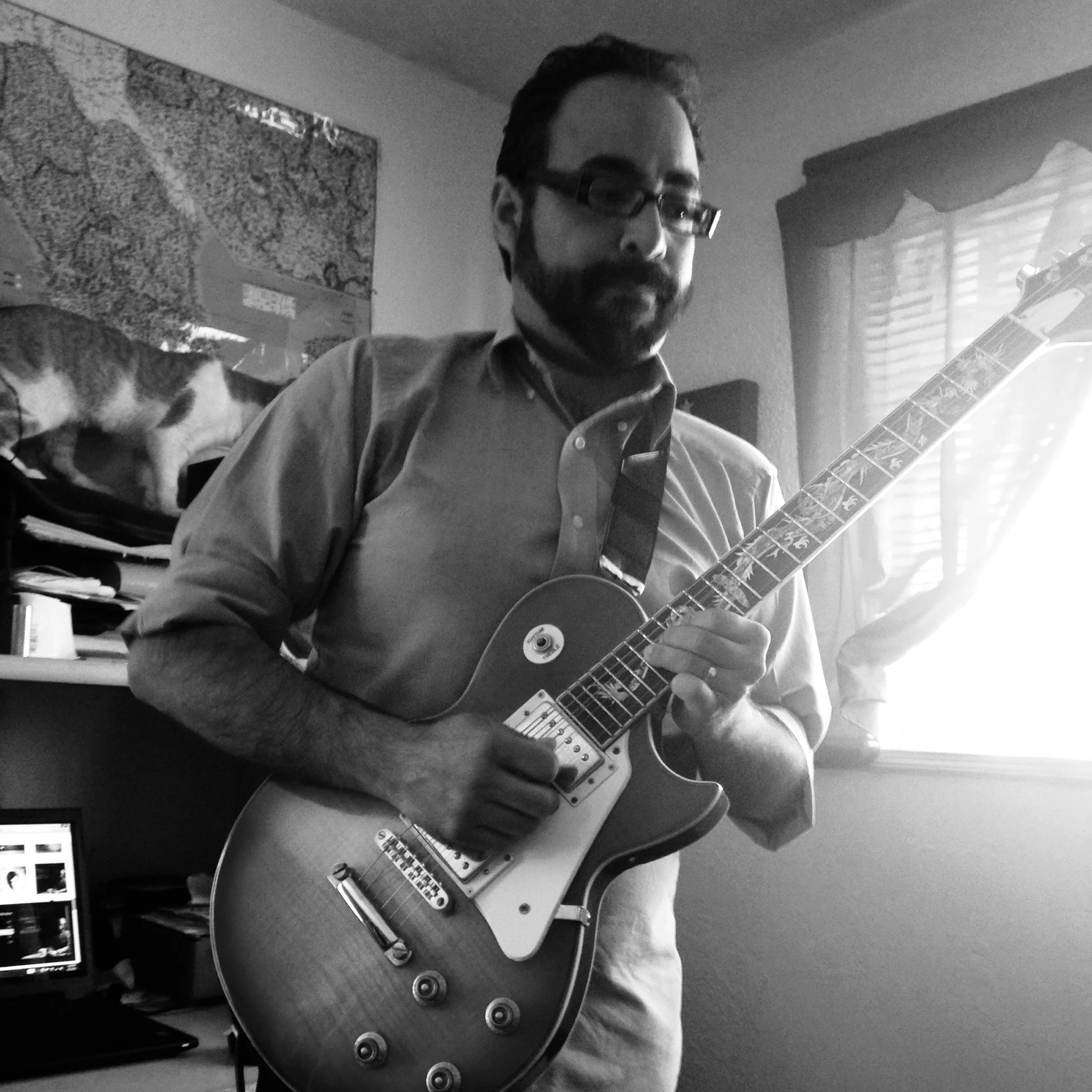 A man with glasses and a beard playing an electric guitar inside a room, with a computer and a cat on a desk in the background, and a window with blinds.