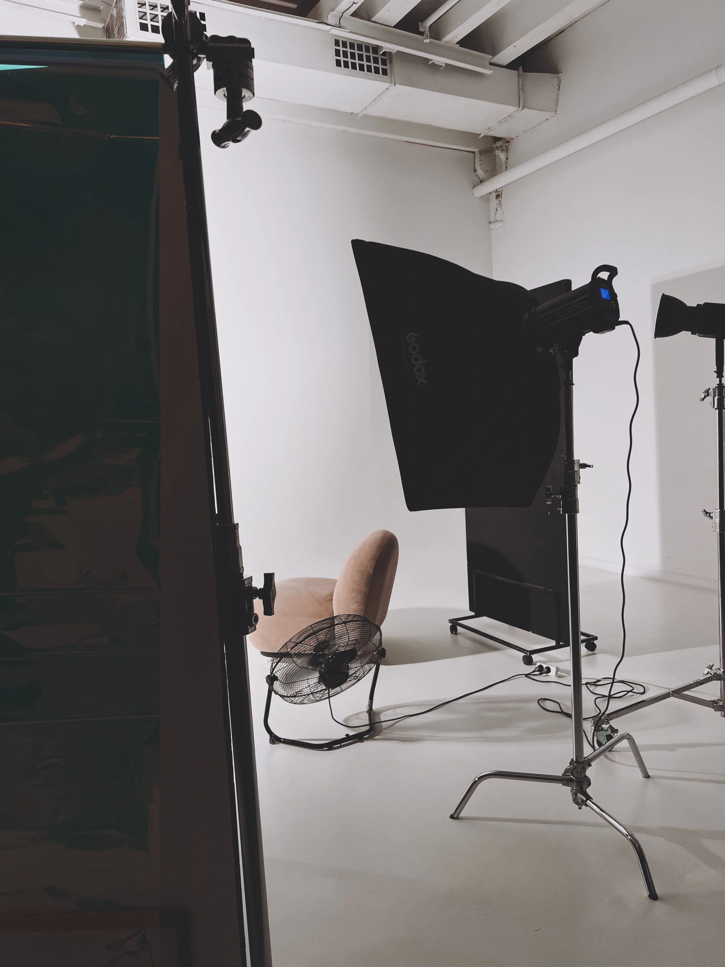 Behind-the-scenes view of a photography studio with lighting equipment, a black reflector, a beige chair, a fan, and a white backdrop.