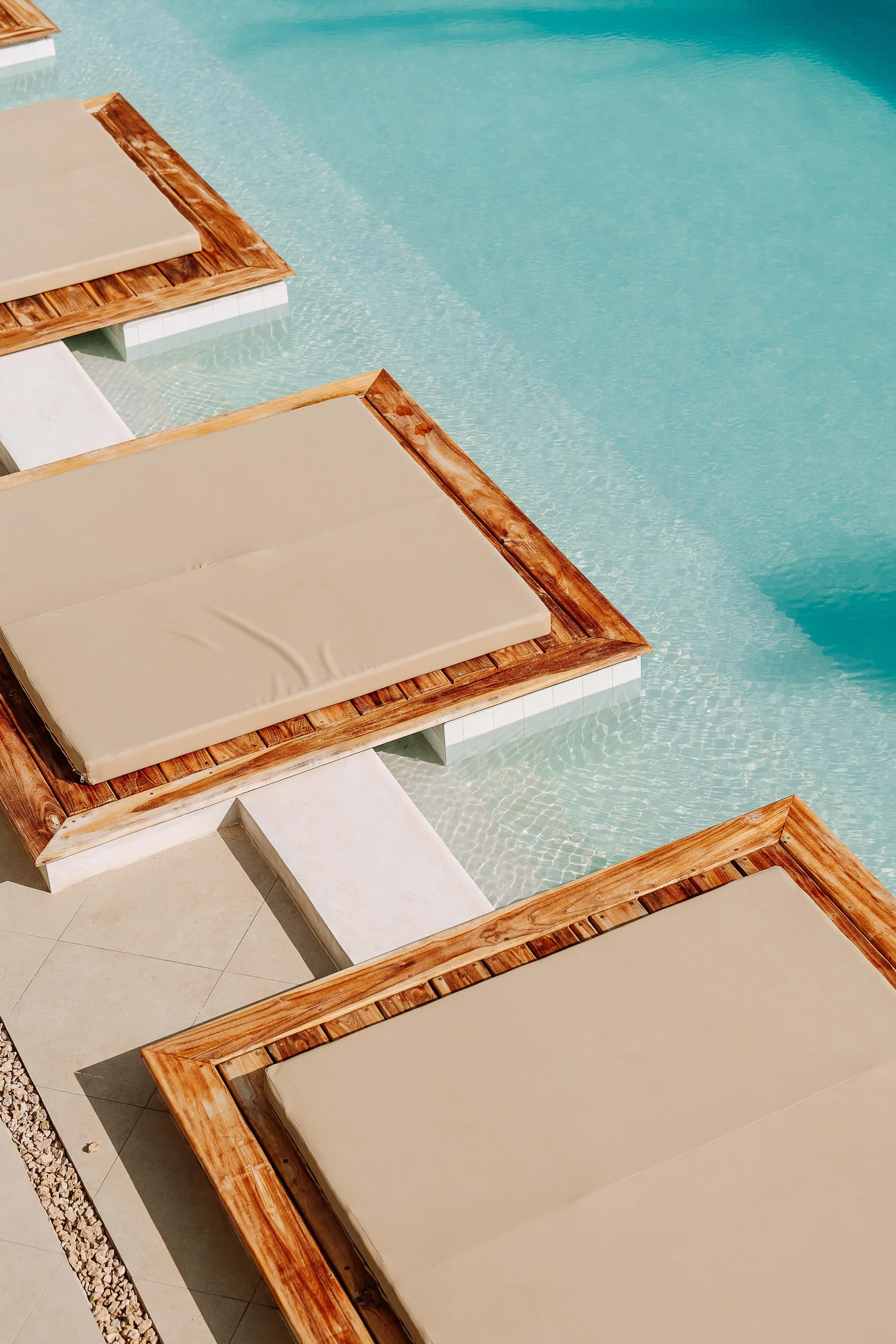 Poolside lounge chairs with beige cushions and wooden frames, overlooking a clear blue swimming pool.