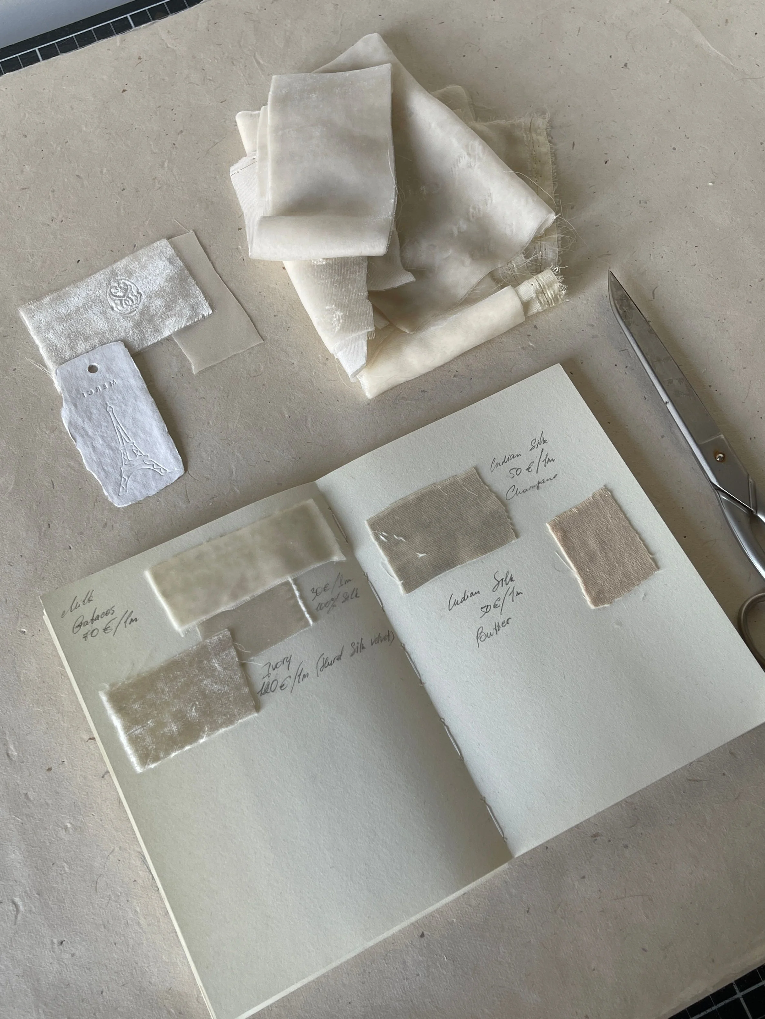 Various fabric swatches and samples arranged on a white sheet of paper or notebook, with handwritten notes describing each fabric. A small piece of fabric with a Paris Eiffel Tower design is visible, along with a pair of scissors on the right side of the image.