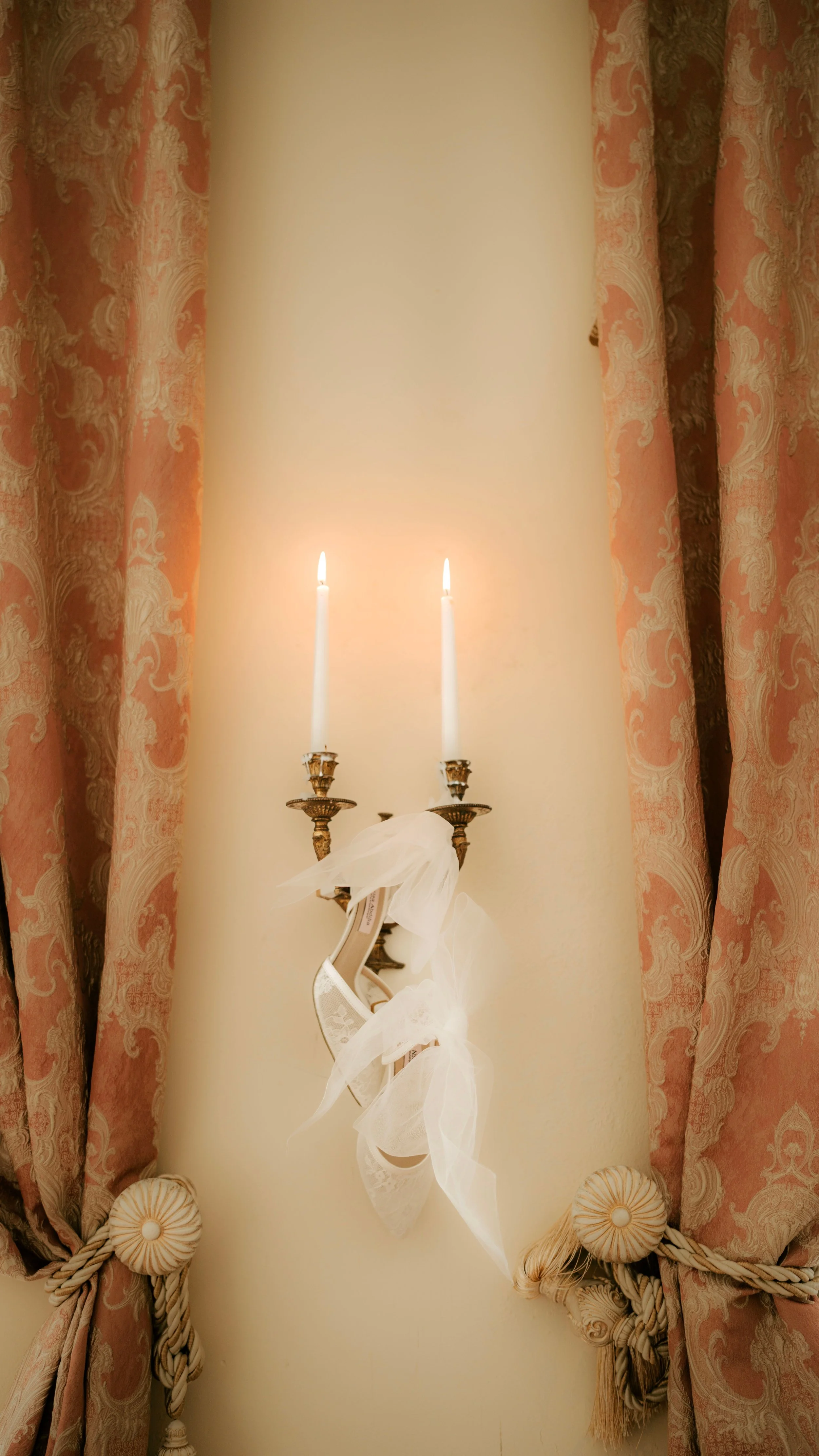 A white wall with pink curtains on either side, featuring gold and cream embellishments and tiebacks, a decorative gold wall sconce with two lit white candles, and white ribbon decorations hanging from the sconce. Bridal Shoes