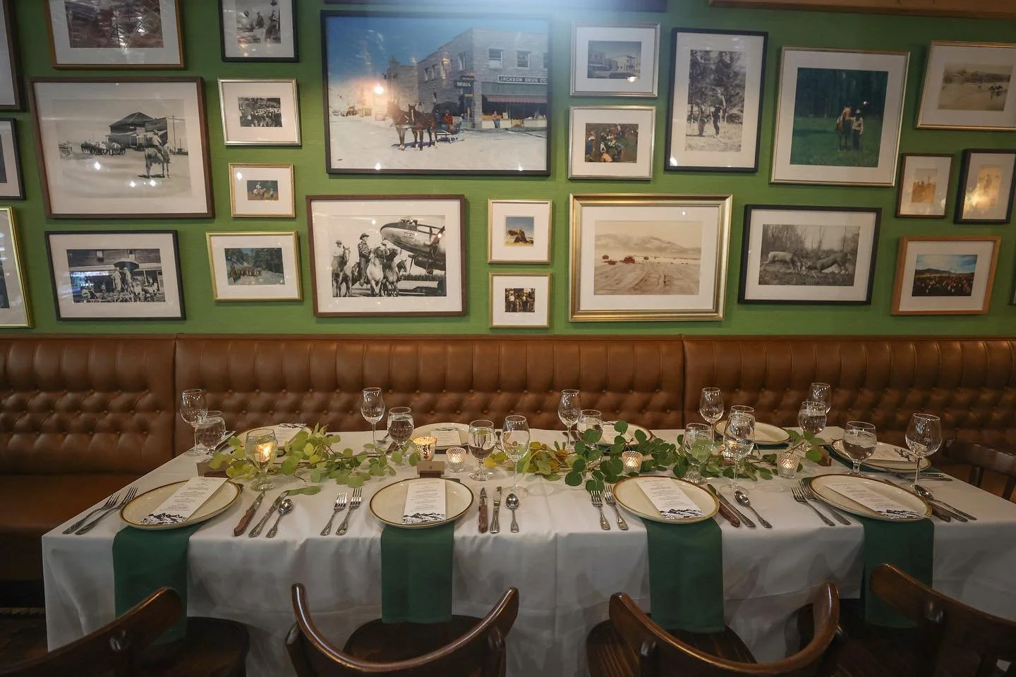 A restaurant table set for a special occasion with a white tablecloth, green runners, and glassware. The wall behind features a green accent wall decorated with a collection of framed photos and artwork, including vintage and modern images.