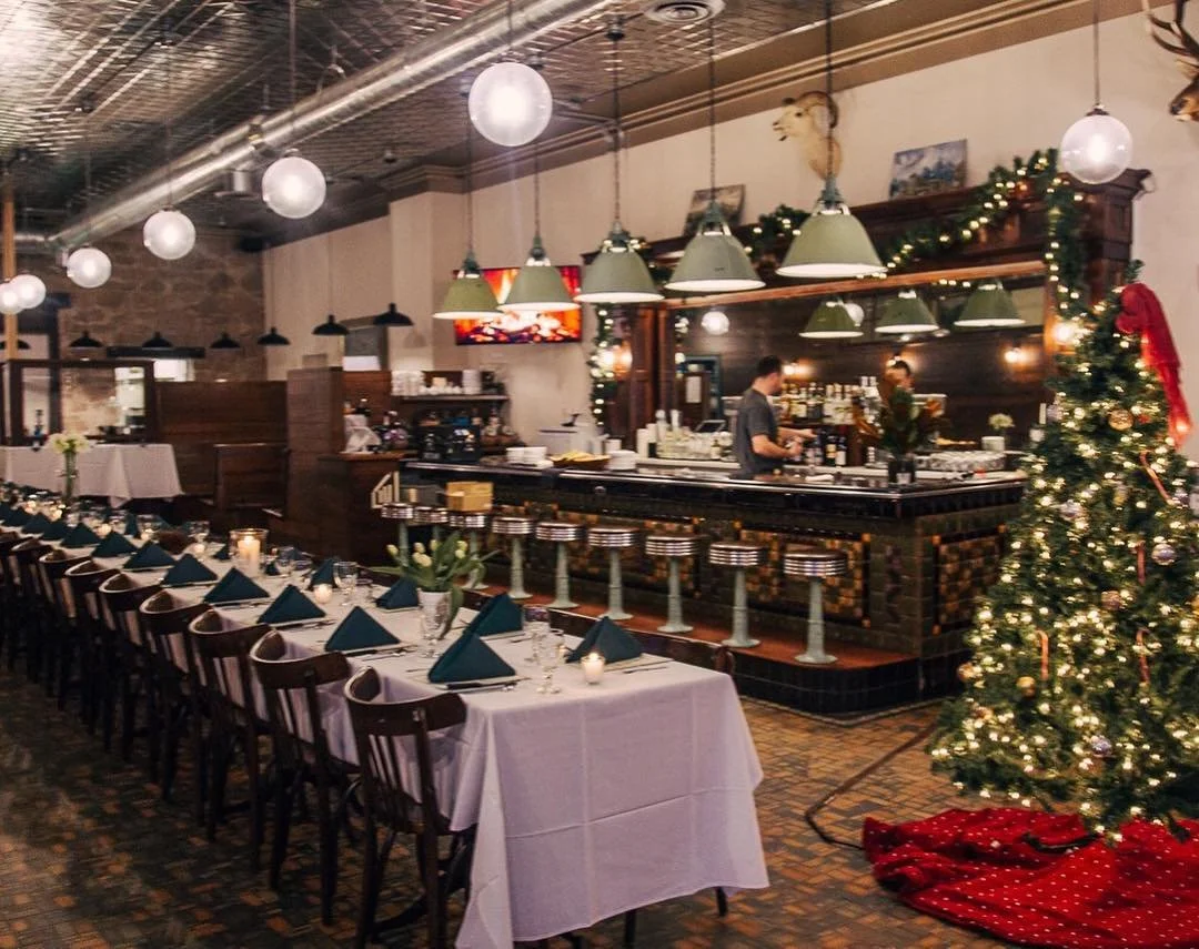 Decorated Christmas tree with lights and ornaments next to a bar area with hanging lights, a long dining table set with candles, napkins, and tableware in a restaurant with festive holiday decor.