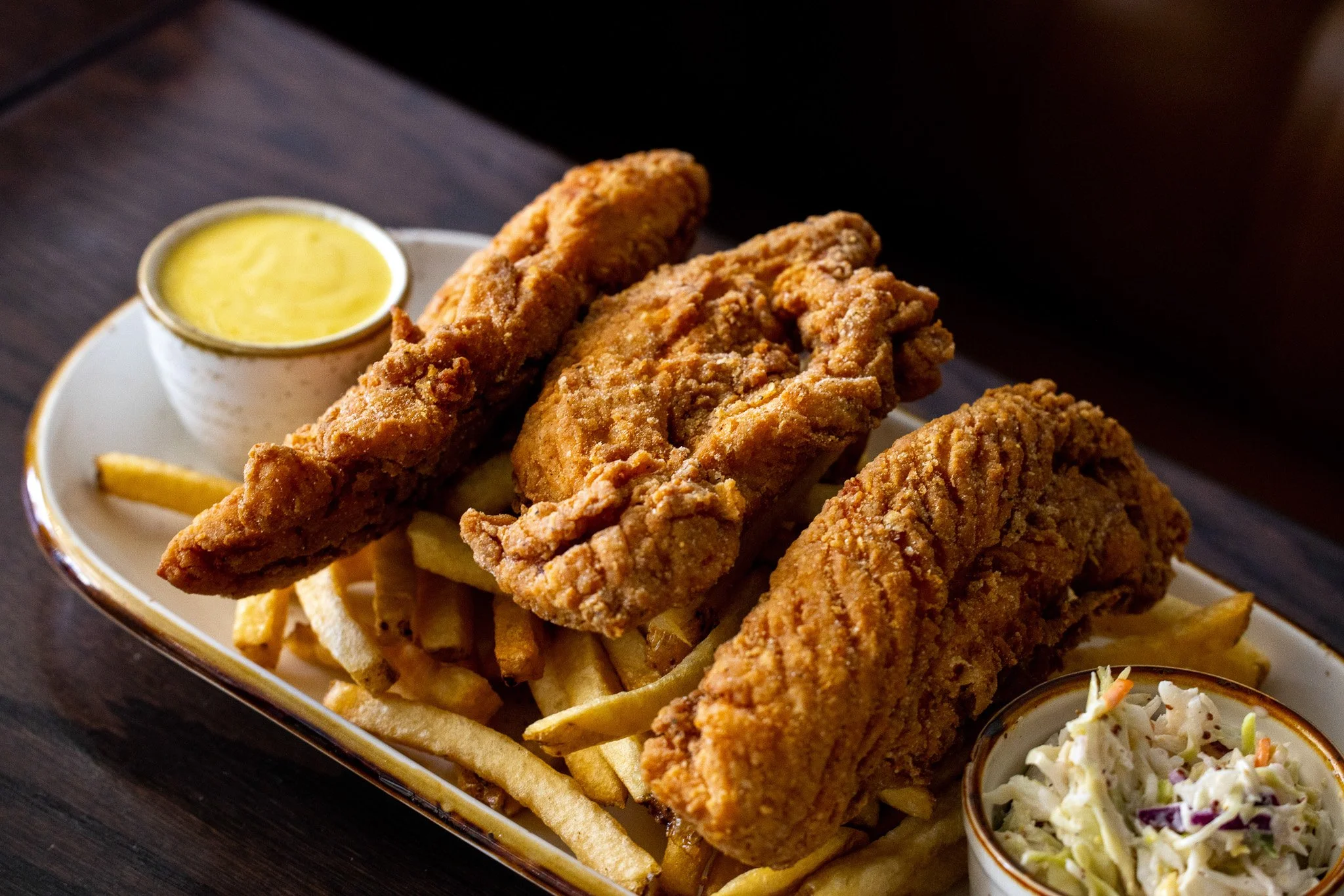 Three pieces of fried fish with French fries, coleslaw, a cup of tartar sauce, and a small bowl of cole slaw on a white platter.