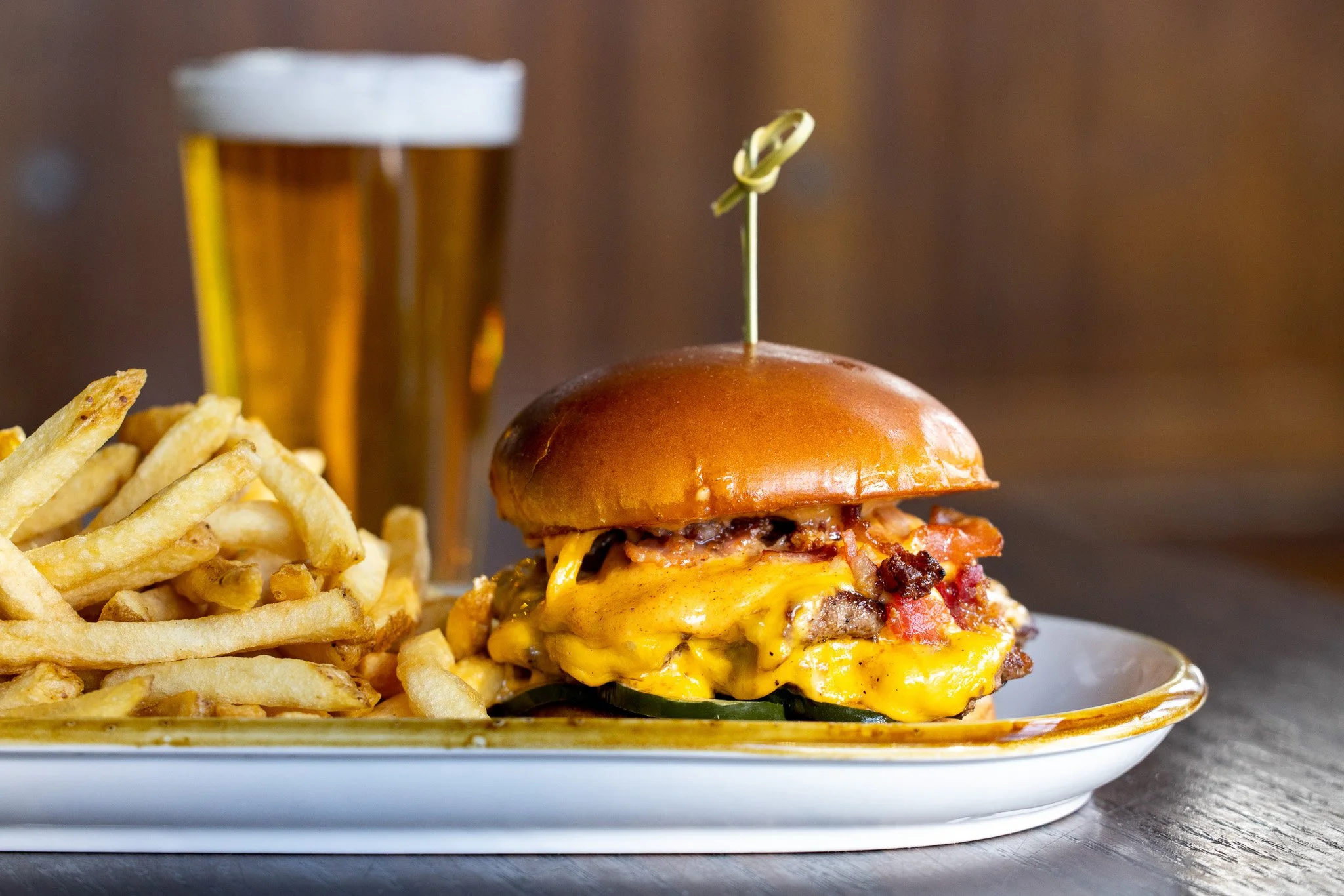 A plate with a cheeseburger topped with bacon and a pick, alongside a side of French fries, and a glass of beer in the background.